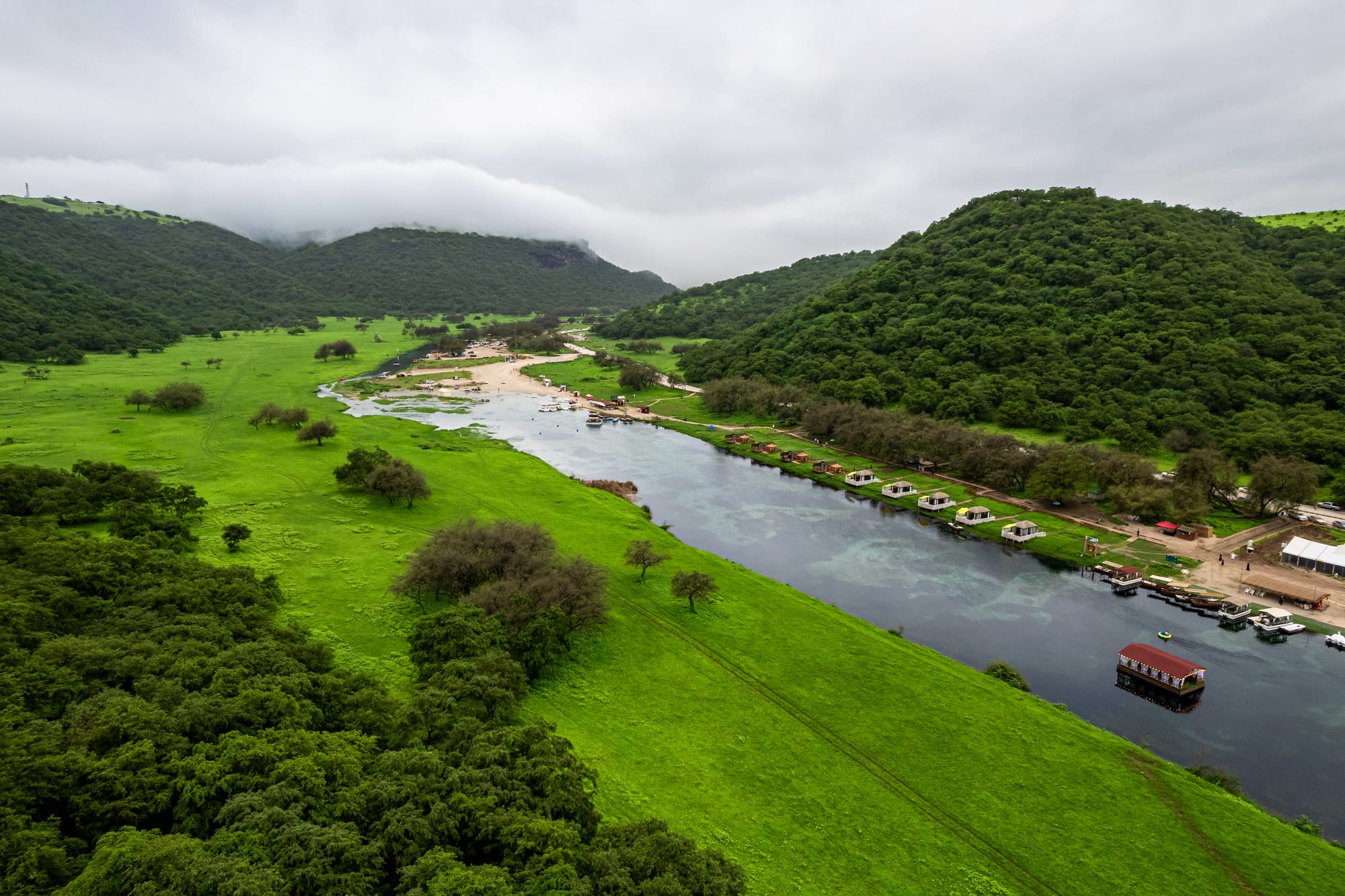 Aerial view of a calm river winding through bright green meadows and wooded hills under low monsoon cloud