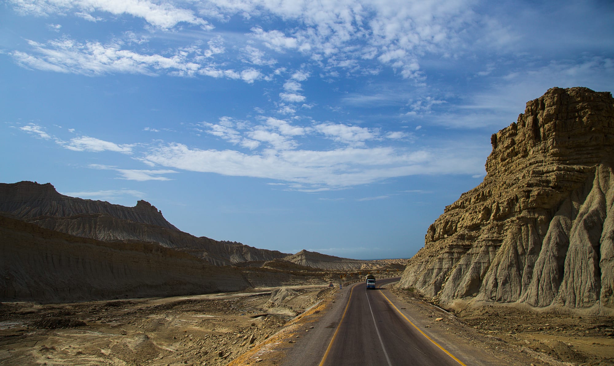 A two-lane road running through a dry valley between eroded, layered sandstone cliffs under a blue sky