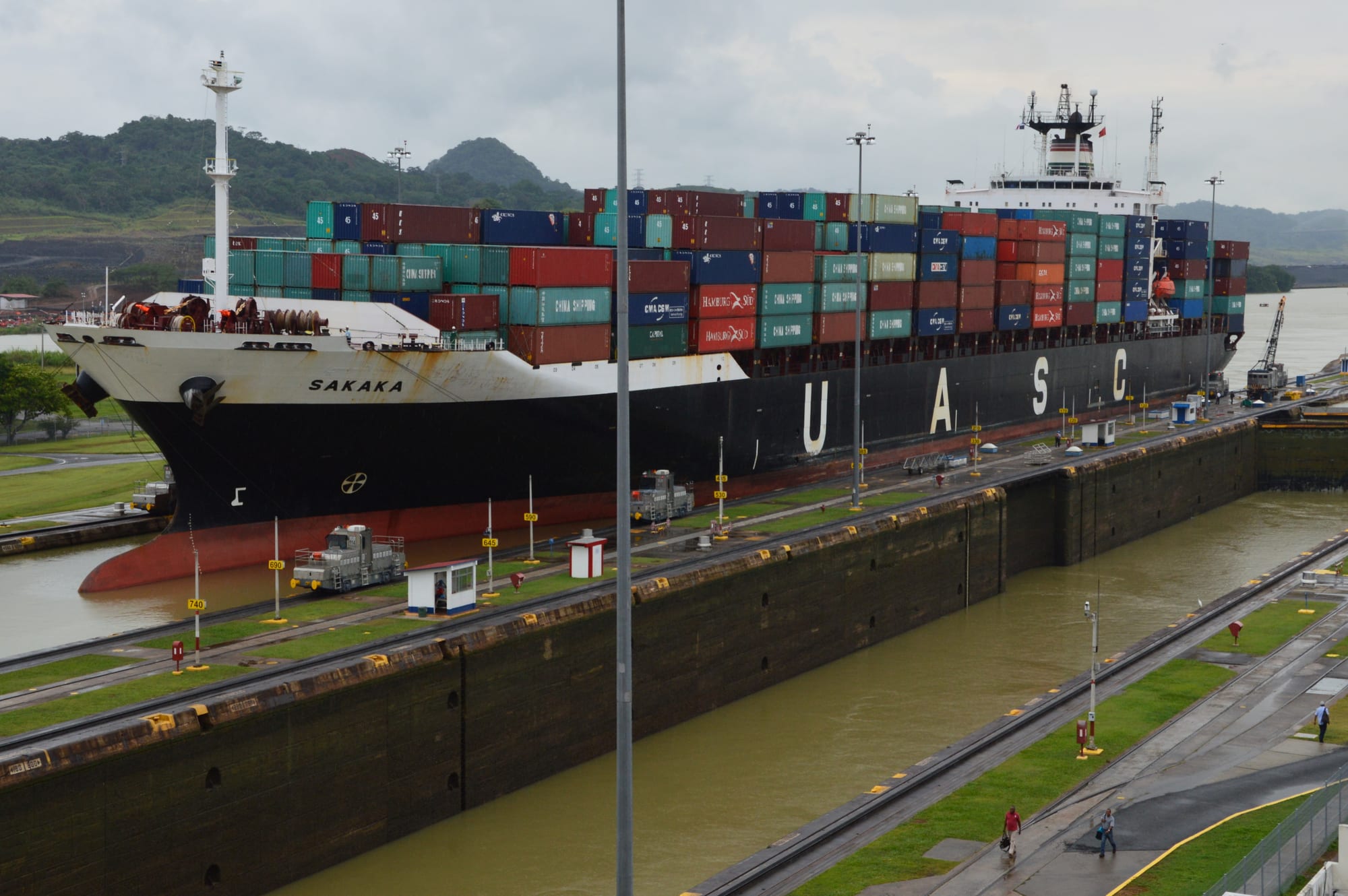 A large container ship stacked with coloured containers rising in a lock chamber, towed by small locomotives beside green hills