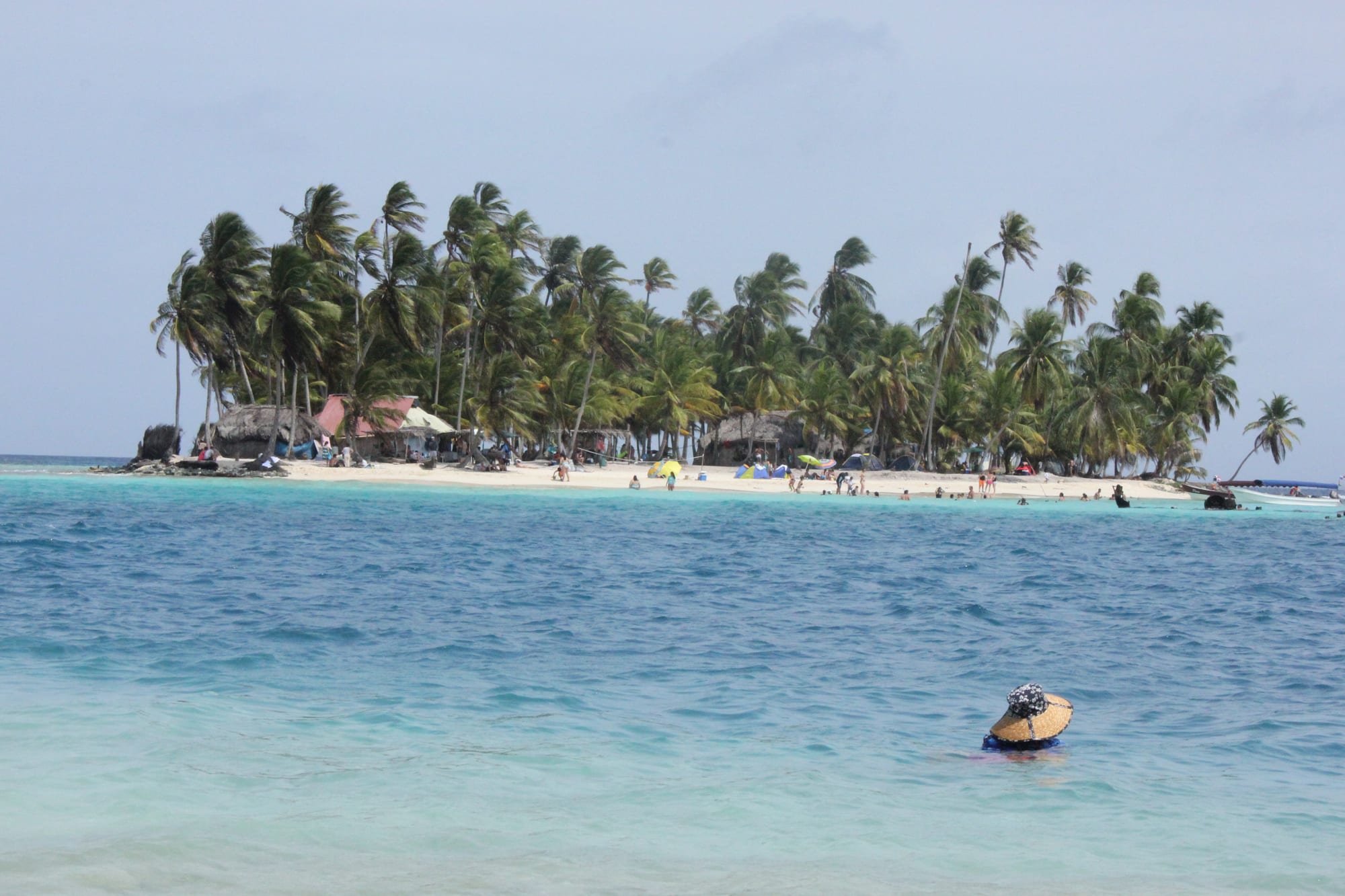 A small island of coconut palms and thatched huts behind a white-sand beach, across clear turquoise water