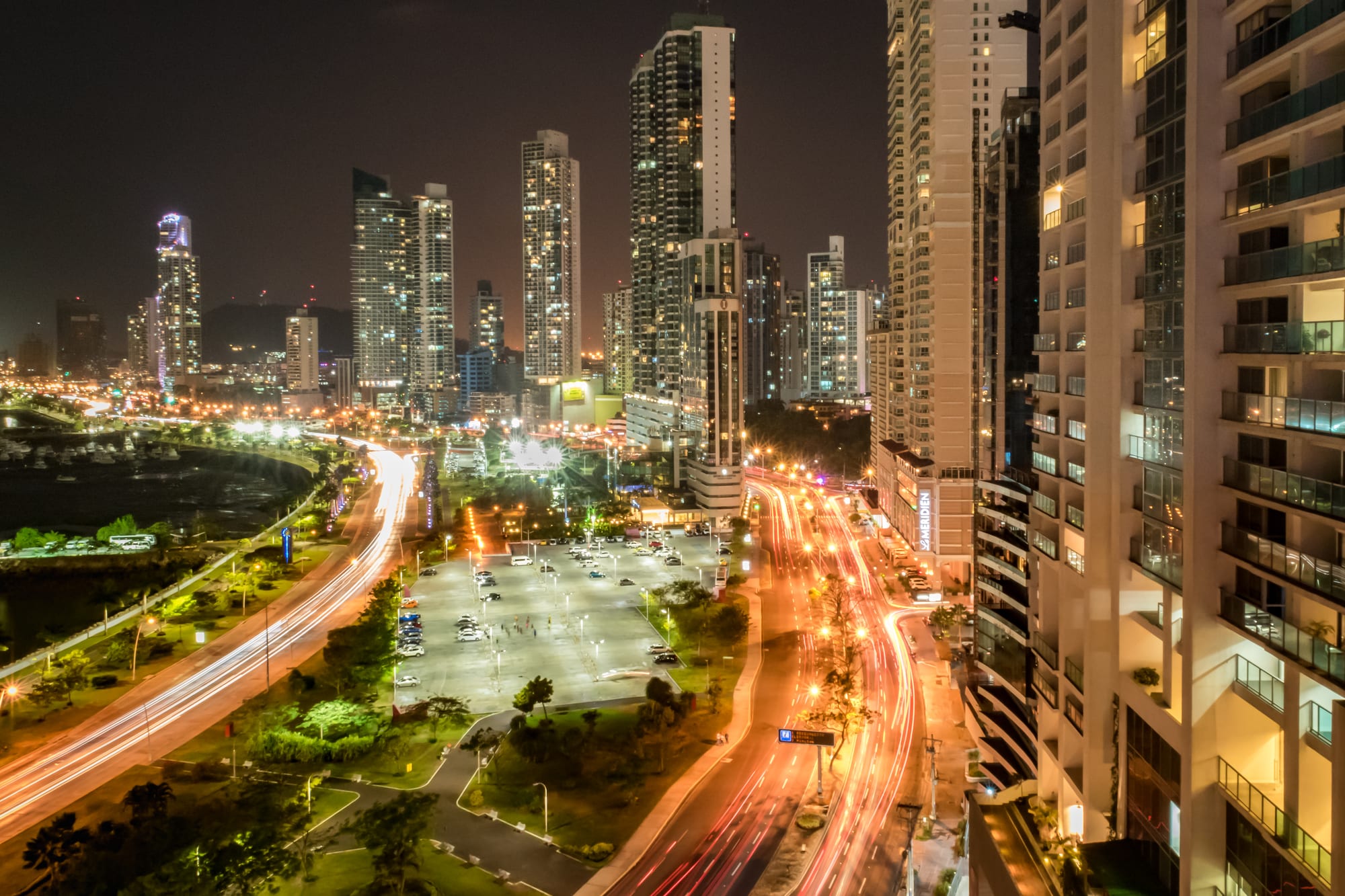 A dense cluster of glass high-rise towers on a curving tropical coastline, green hills and open Pacific water beyond