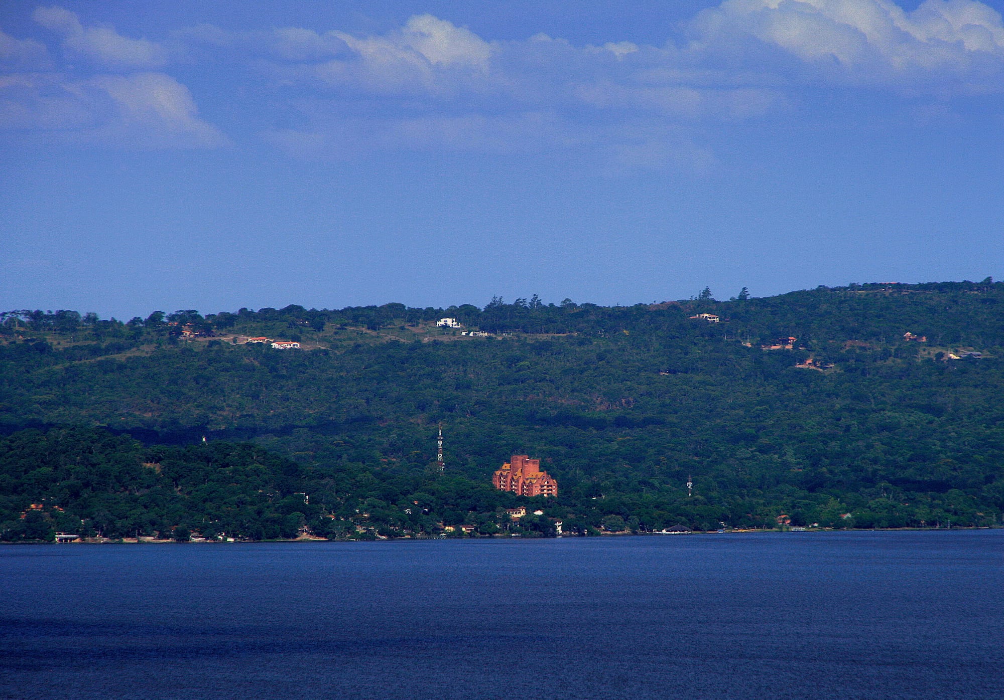 Dark blue lake water in front of a long wooded hillside dotted with houses and a large red-brick building near the shore