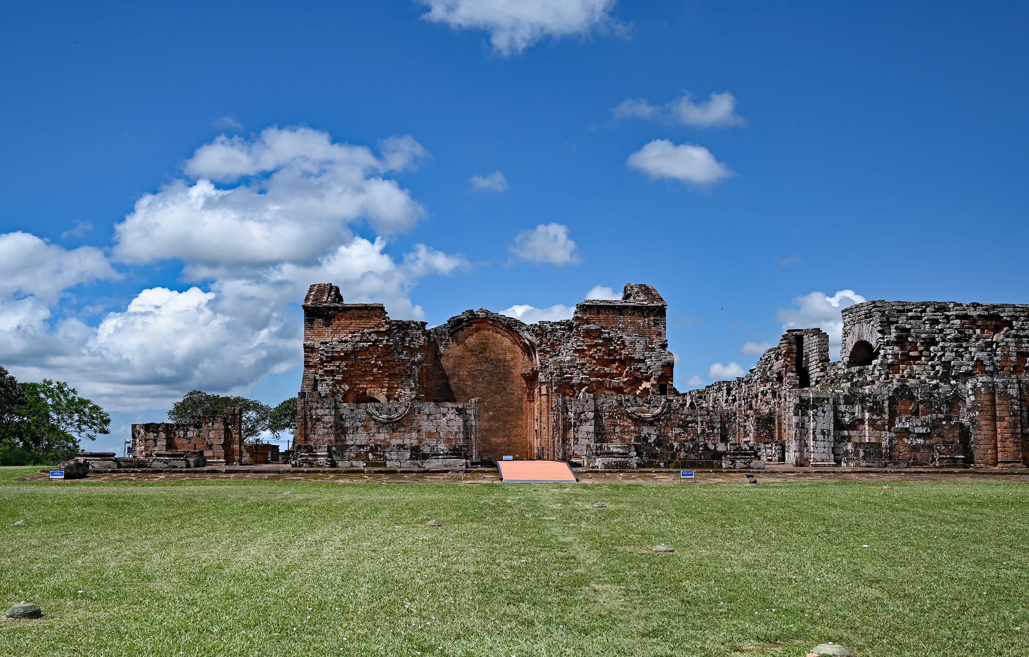 The roofless red-sandstone walls and arches of a large baroque mission church standing on a grassy plain under a blue sky
