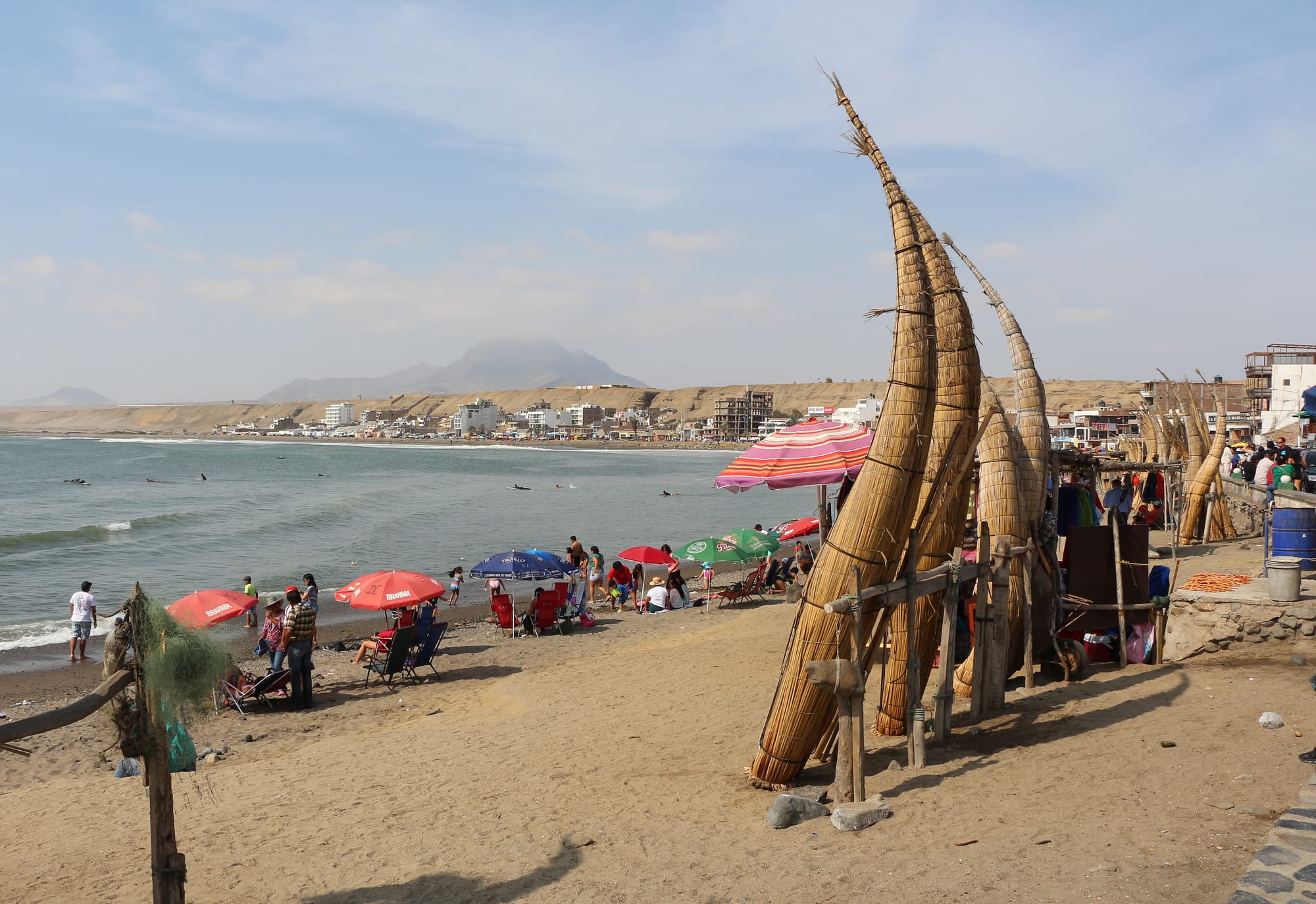Tall reed fishing boats propped upright on a sandy beach, with umbrellas, surfers in the sea and a low town beyond