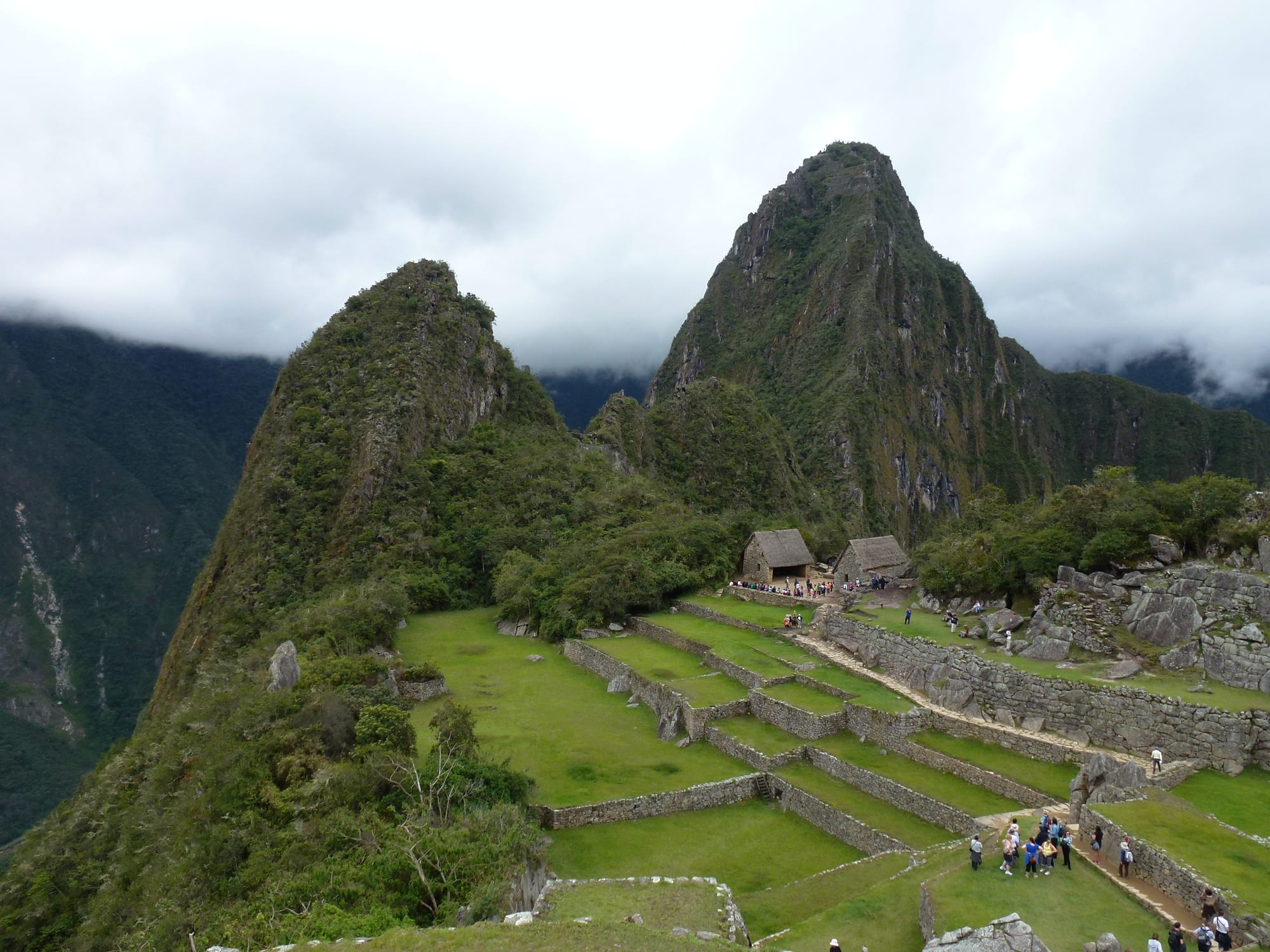 Stone Inca ruins and green agricultural terraces on a high mountain saddle, with the steep peak of Huayna Picchu rising behind under a clear sky