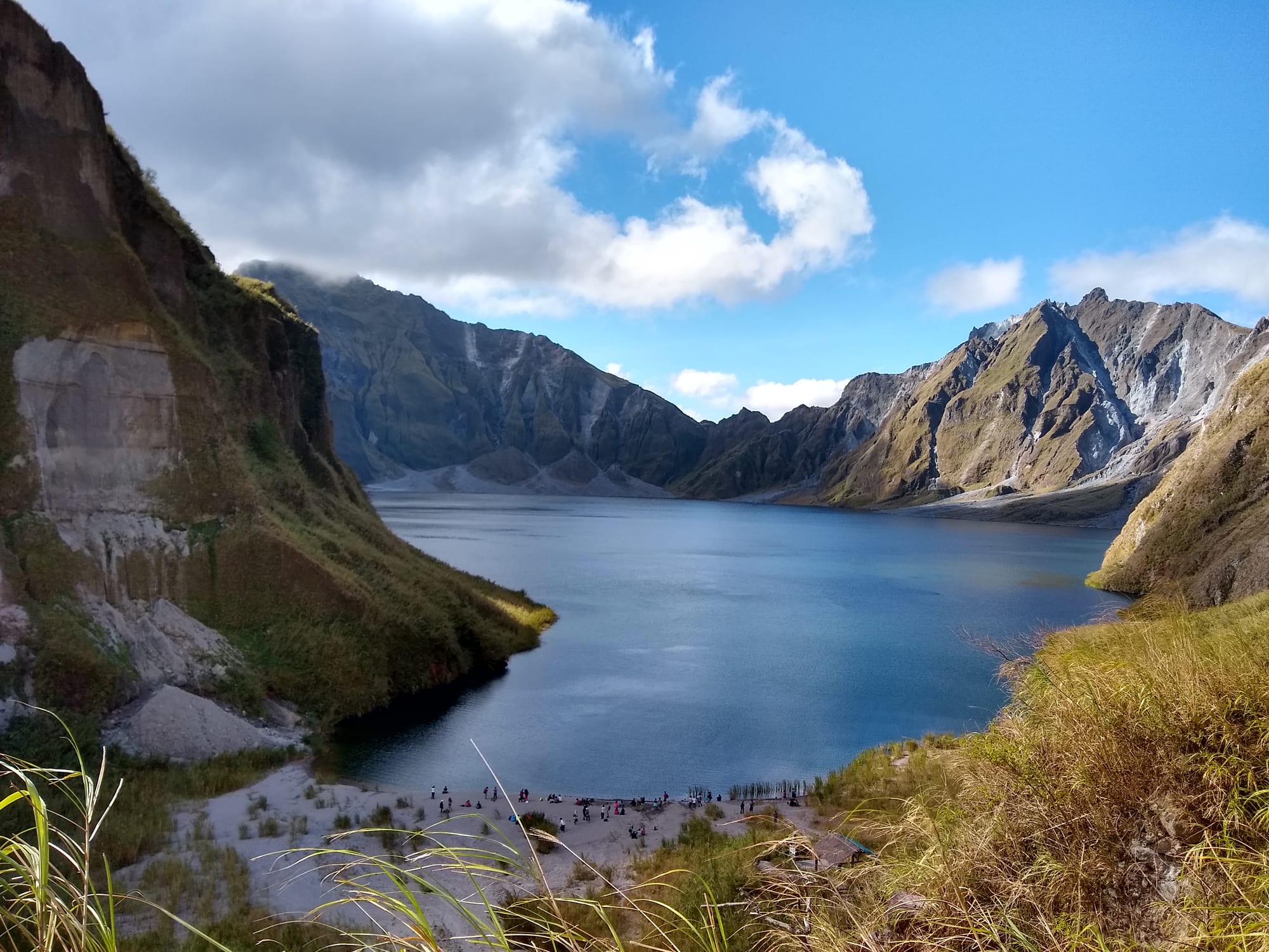 Blue crater lake ringed by steep grey-green volcanic cliffs, with grassy slopes and a pale sandy shore in the foreground