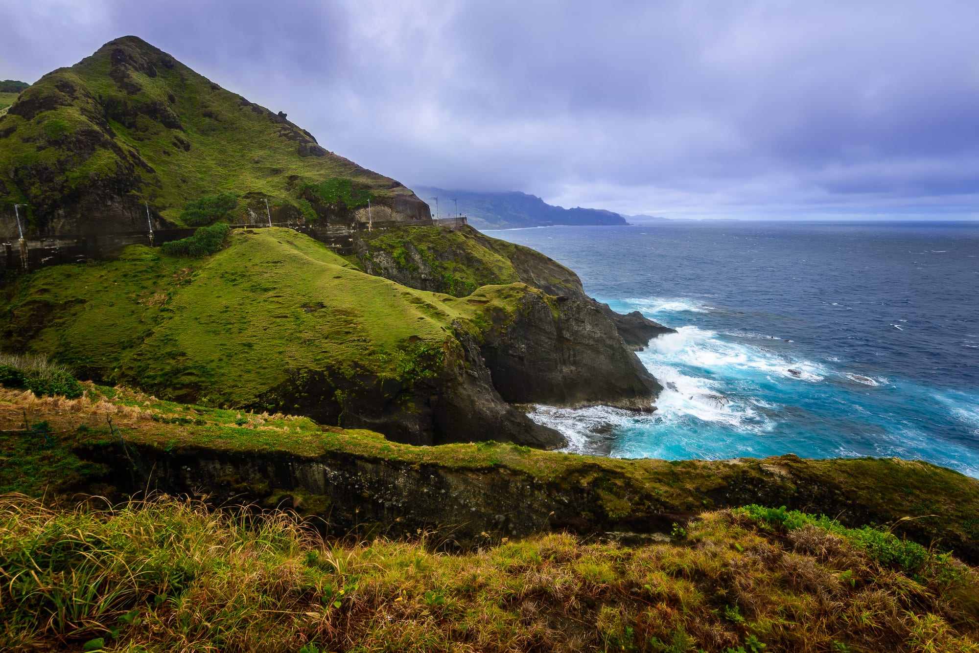 Green grassy hills ending in dark rocky cliffs above a turquoise sea with white surf, under a heavy grey sky