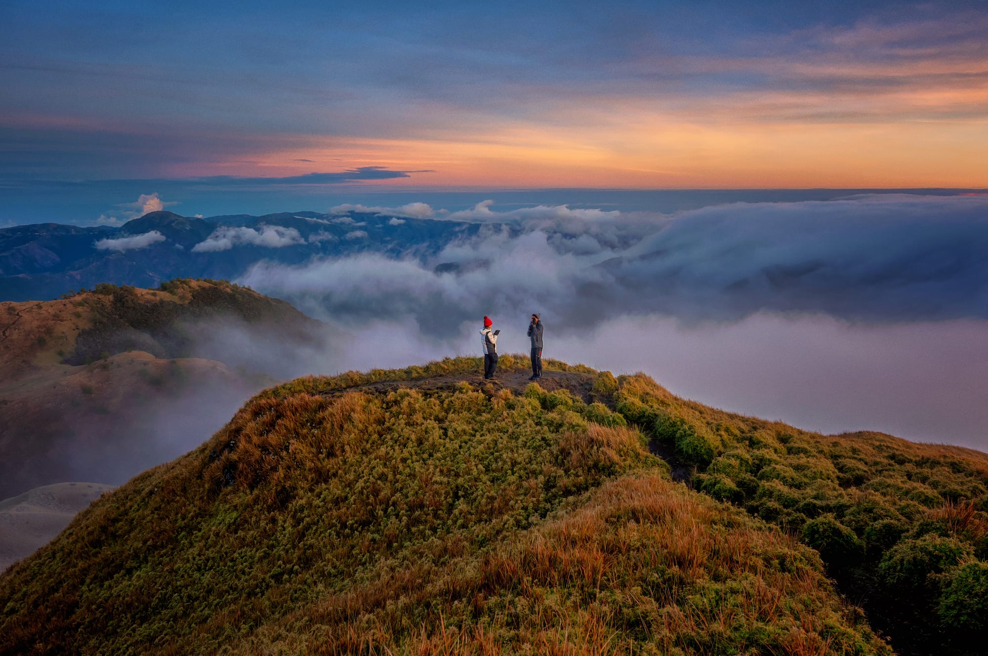 Two hikers stand on a grassy summit ridge above a rolling sea of clouds, with blue mountain ranges behind and an orange and pink sunrise sky