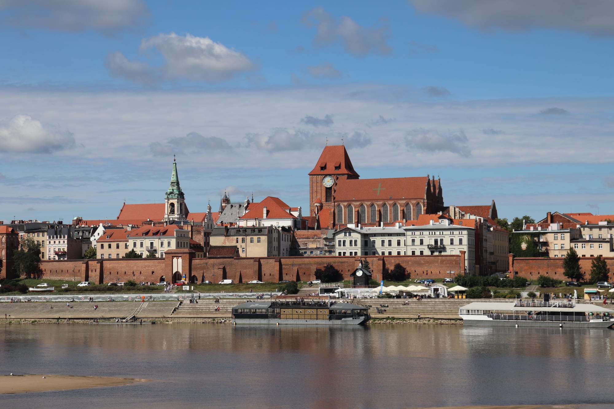 A red-brick Gothic old town with a large church, a green spire and a long defensive wall, seen across a wide river with moored boats