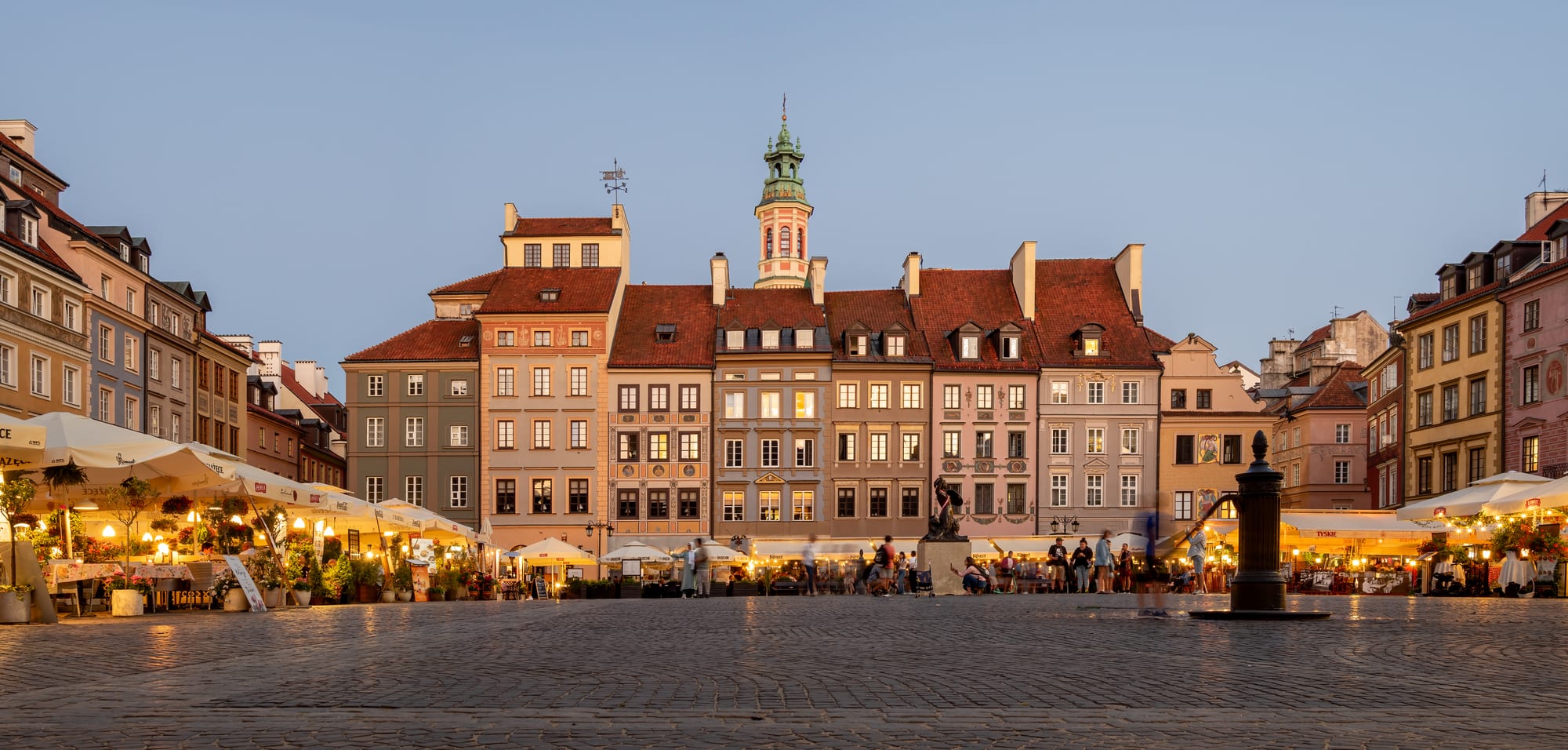 Colourful tall townhouses with red roofs and a small tower lining Warsaw's cobbled Old Town square at dusk