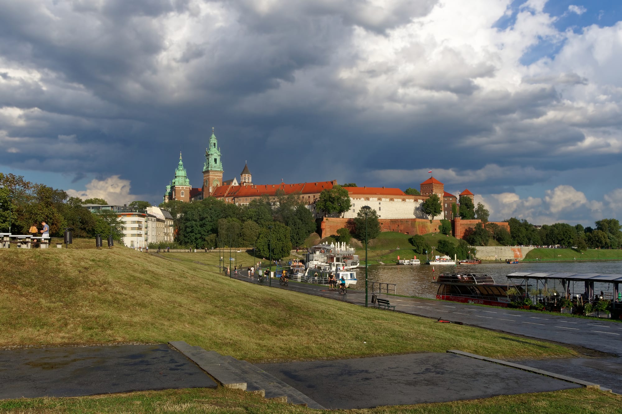 A hilltop complex of Renaissance castle buildings and a cathedral with copper domes rising above a green riverbank and the wide Vistula