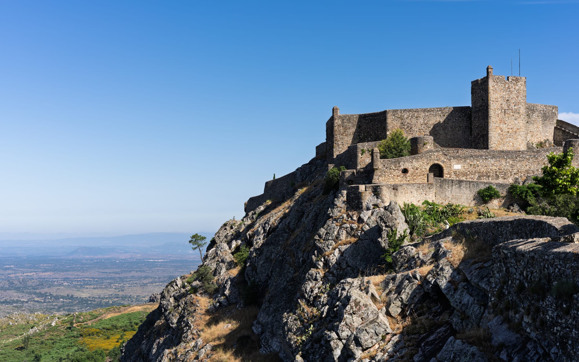 Stone castle walls and a square keep on top of a rocky crag, with a wide hazy plain stretching away below