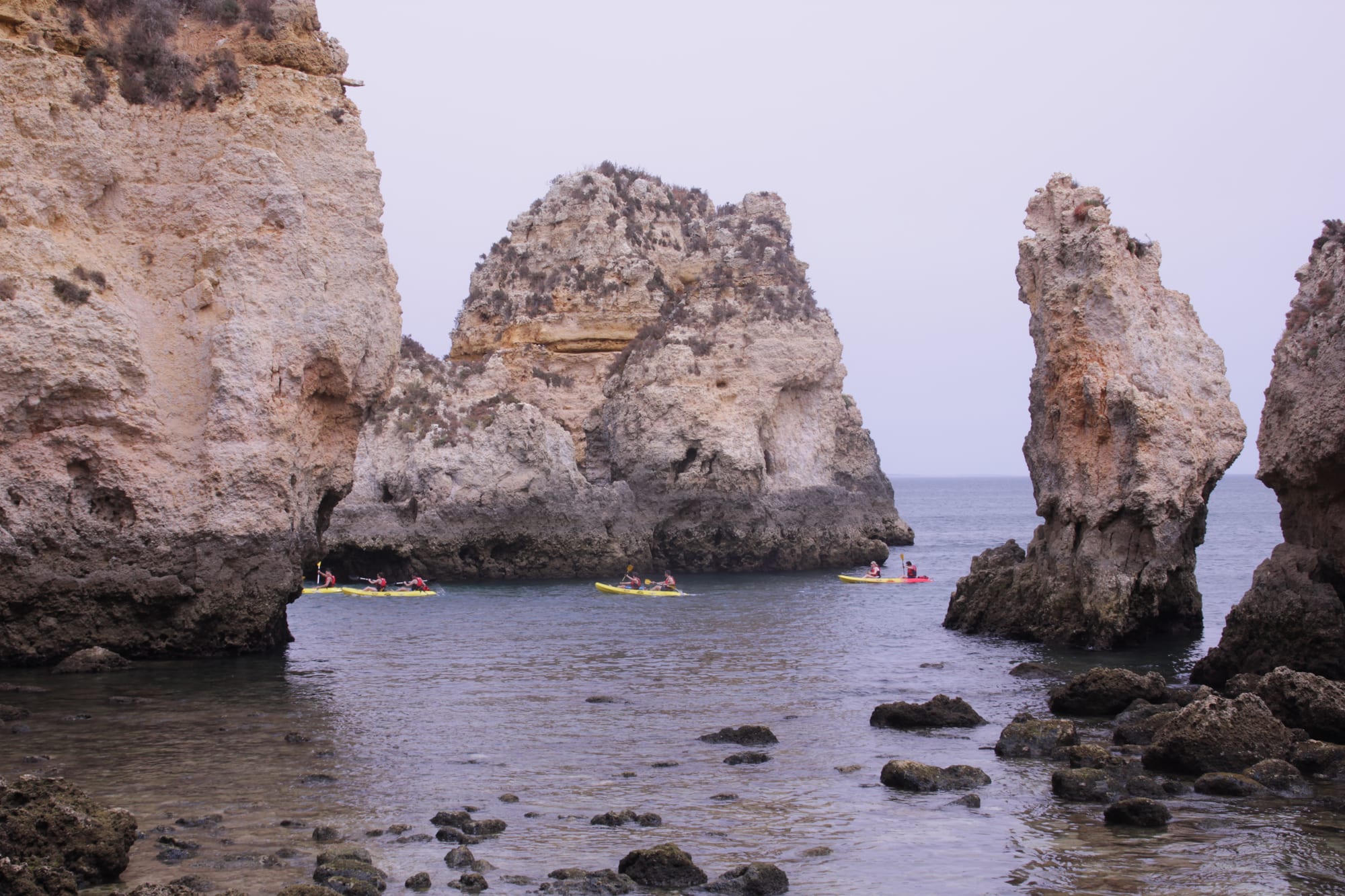 Golden limestone stacks rising from calm sea water, with a line of yellow kayaks passing between the rocks