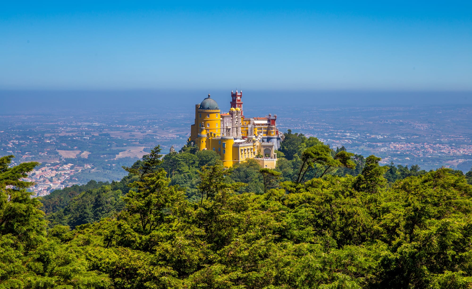A yellow and red palace with turrets and a dome rising above green forest, with a wide plain stretching to the horizon