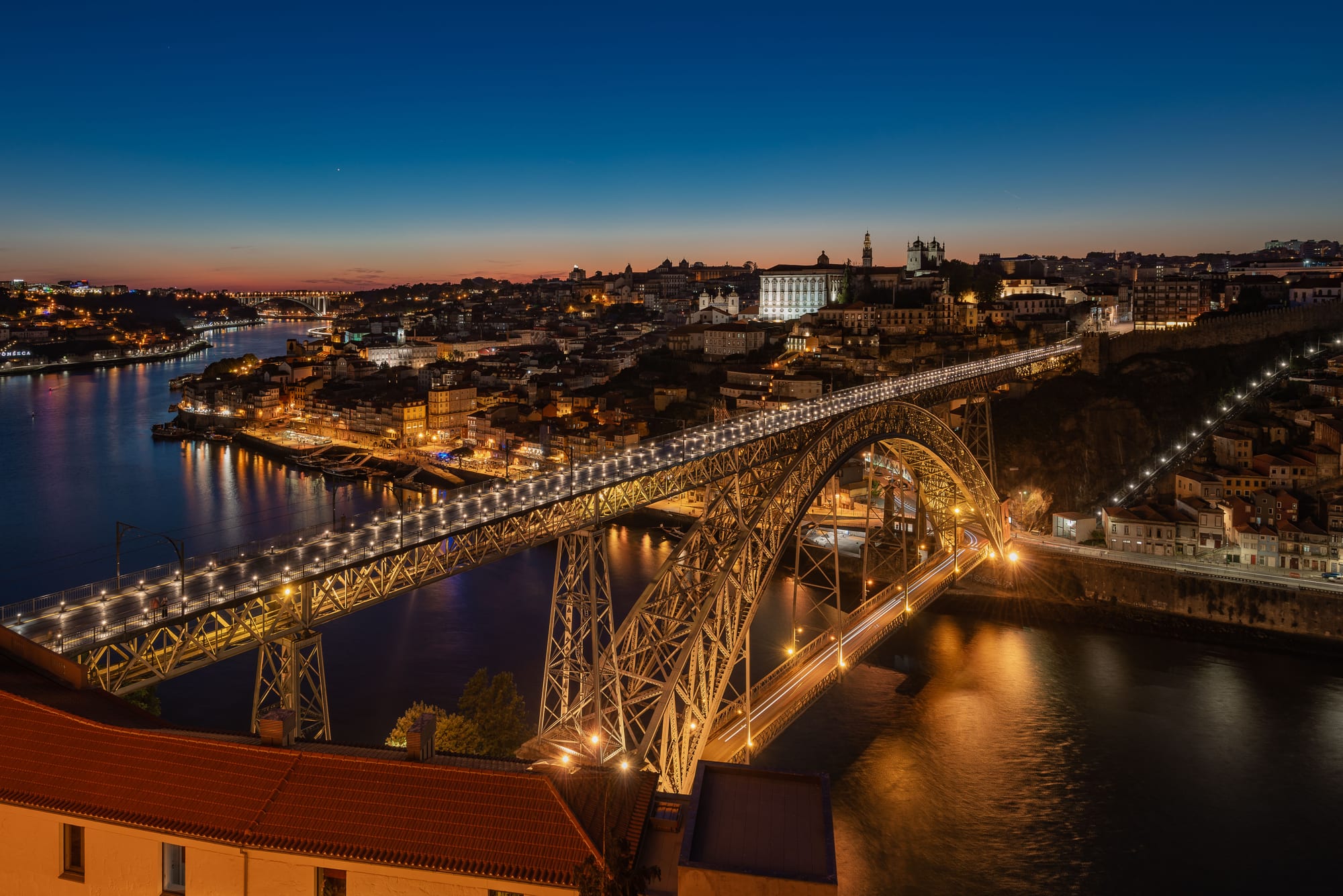 A double-deck iron arch bridge lit in gold spans a dark river at twilight, with the lights of a hillside old town behind and an orange glow on the horizon under a deep blue sky