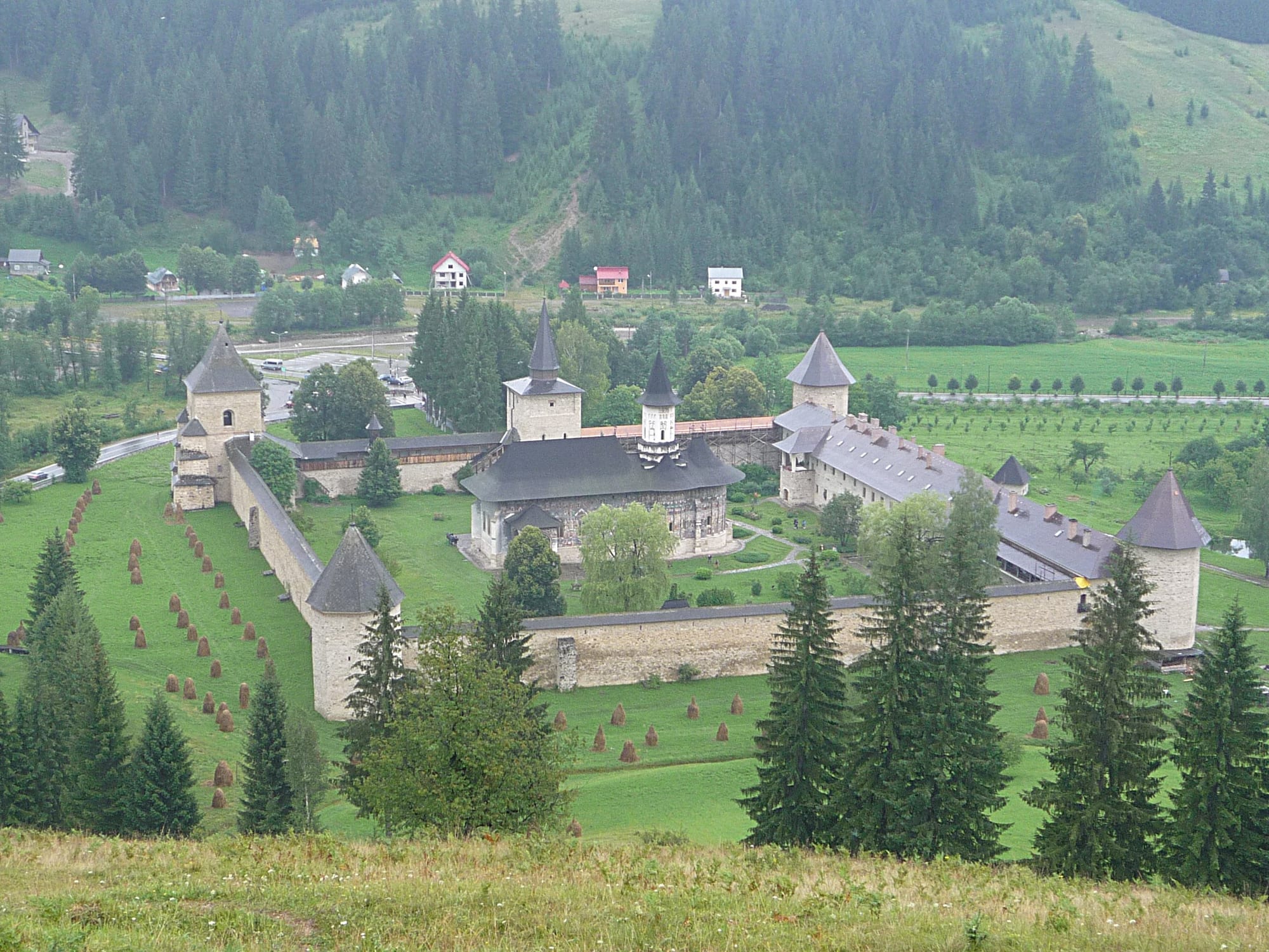 A walled monastery with corner towers and a painted church in a green valley, with haystacks and wooded hills around