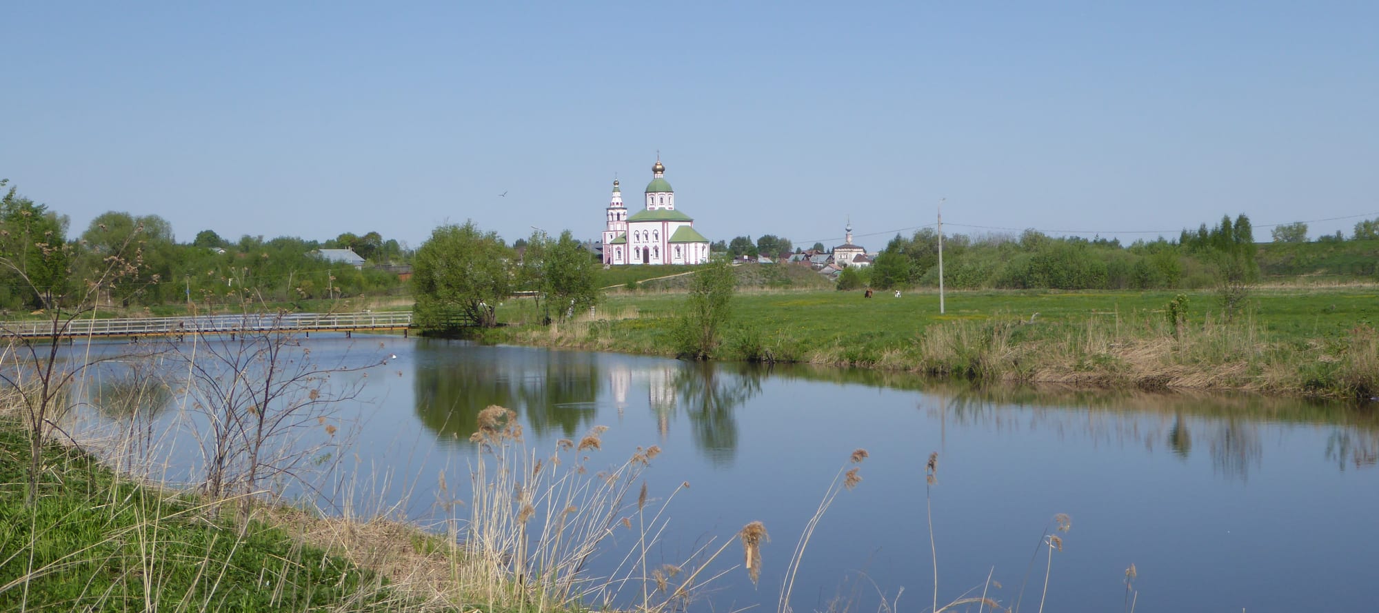 A white church with green domes above water meadows and the still Kamenka river, reflected under a clear blue sky