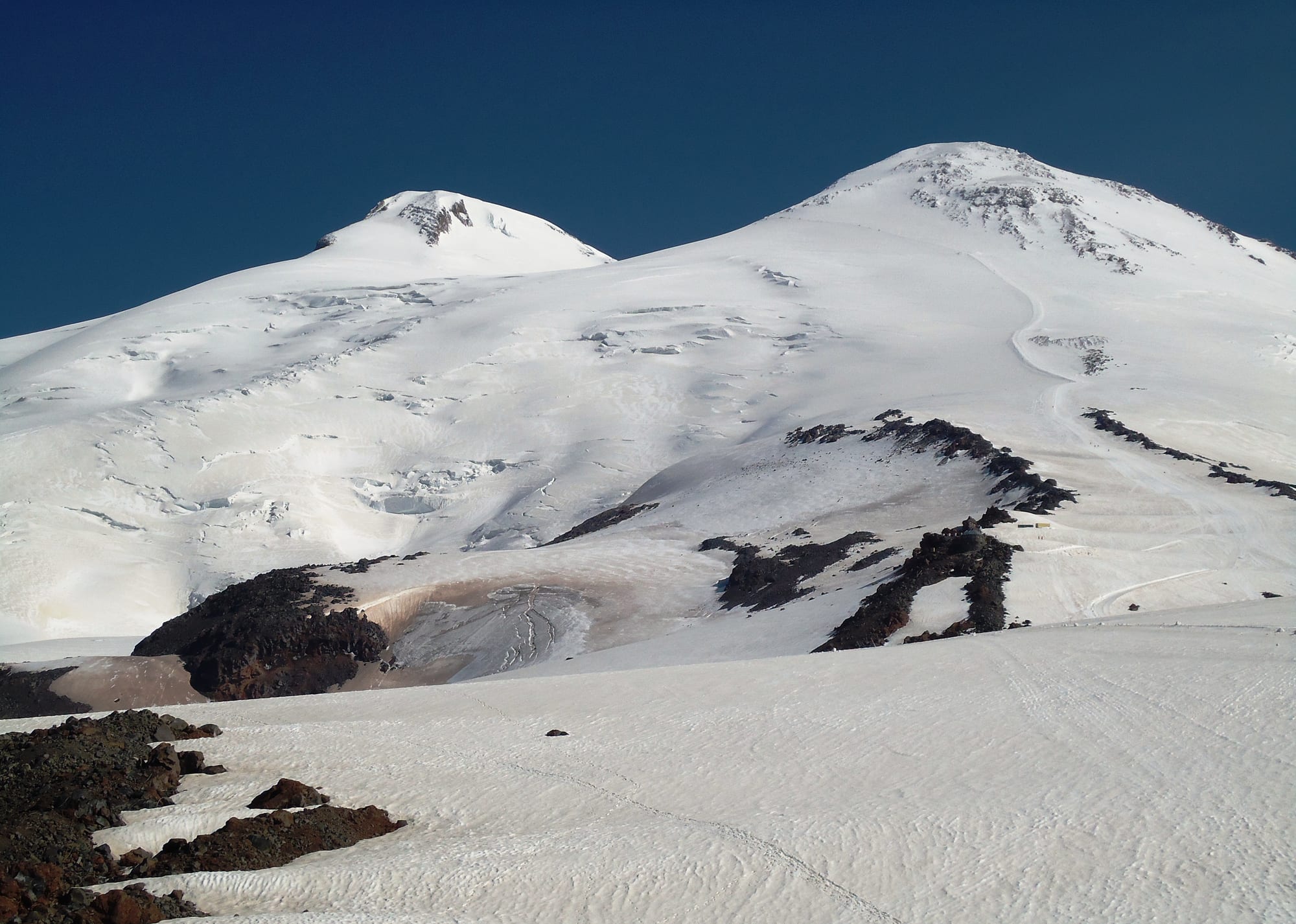 The two snow-covered summits of Mount Elbrus rising above glaciers and dark volcanic rock under a deep blue sky