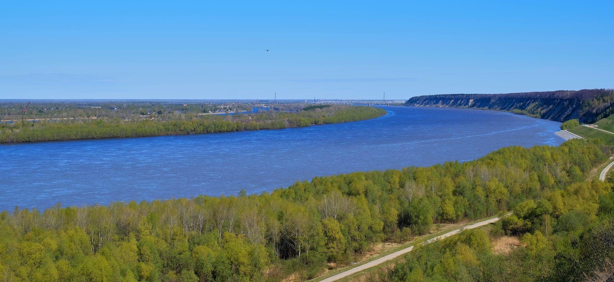 A broad blue river curving between green spring woods and a high bank under a clear sky, with a bridge in the distance