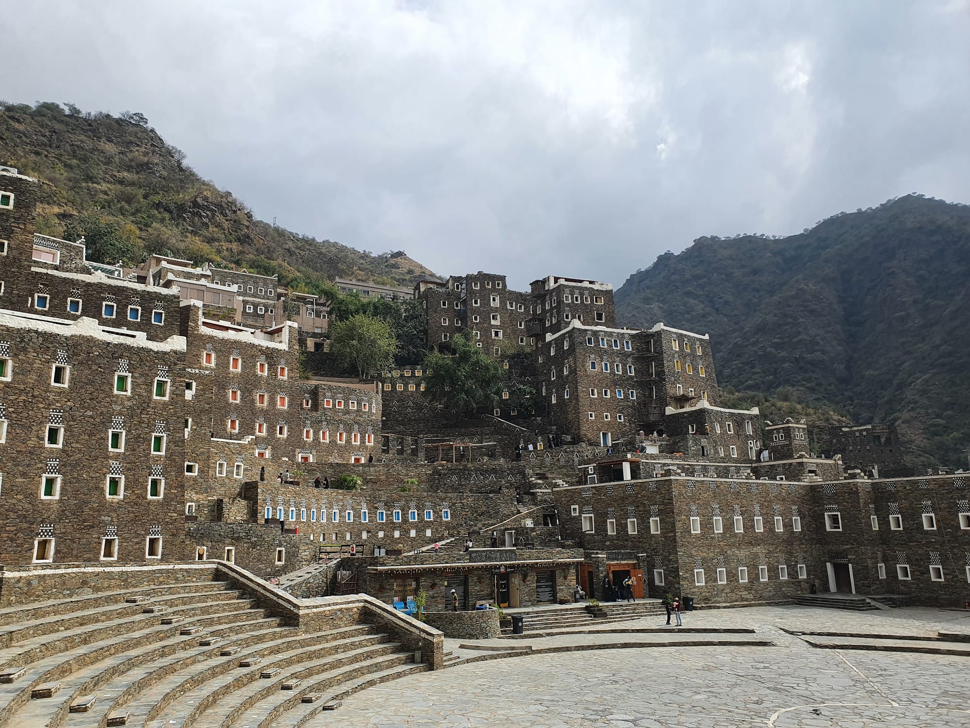 Tall stone tower-houses with white-framed windows stacked up a hillside below green mountains under a cloudy sky