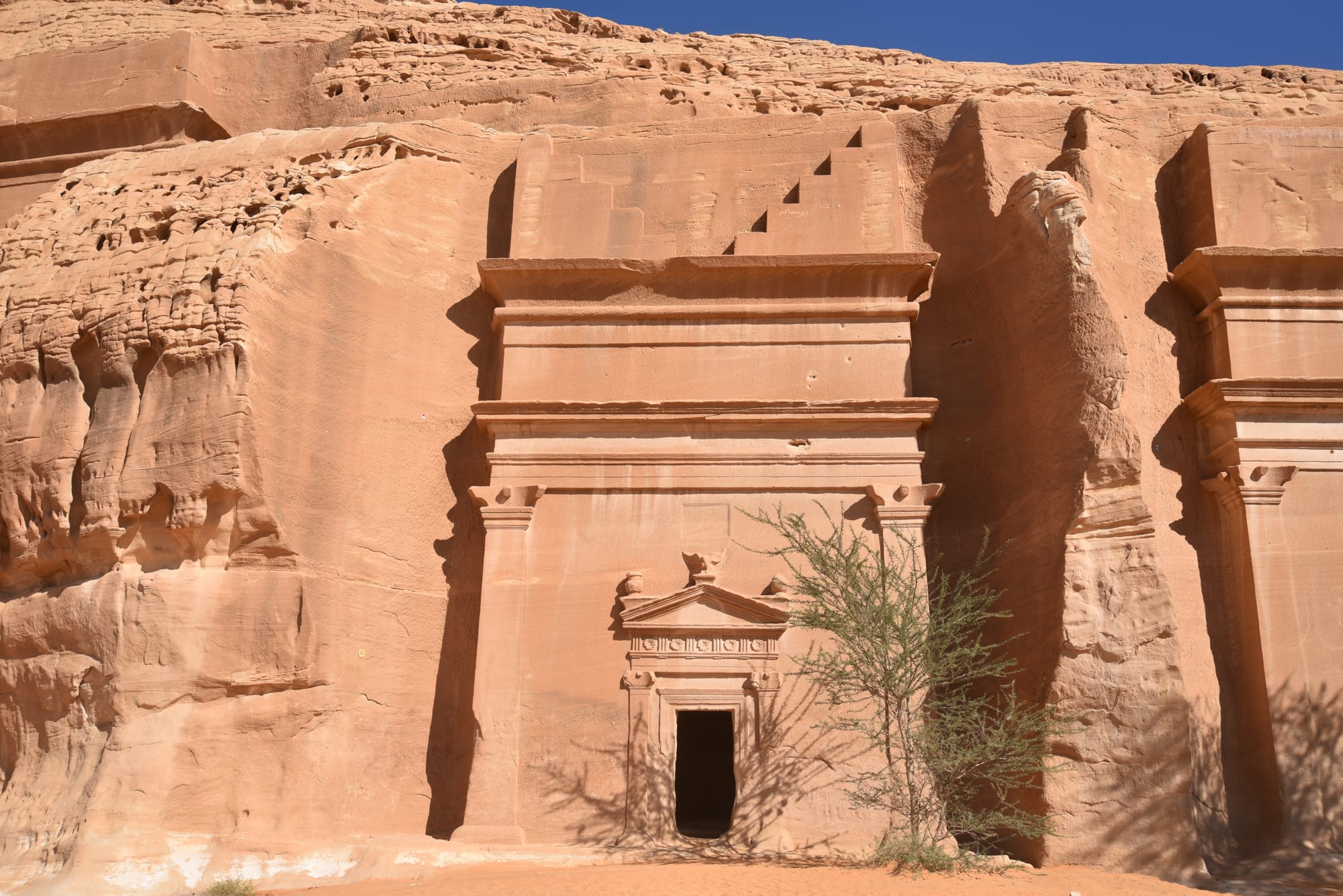 A Nabataean tomb facade with a stepped crown and carved doorway cut into a pink sandstone cliff, a small tree in front