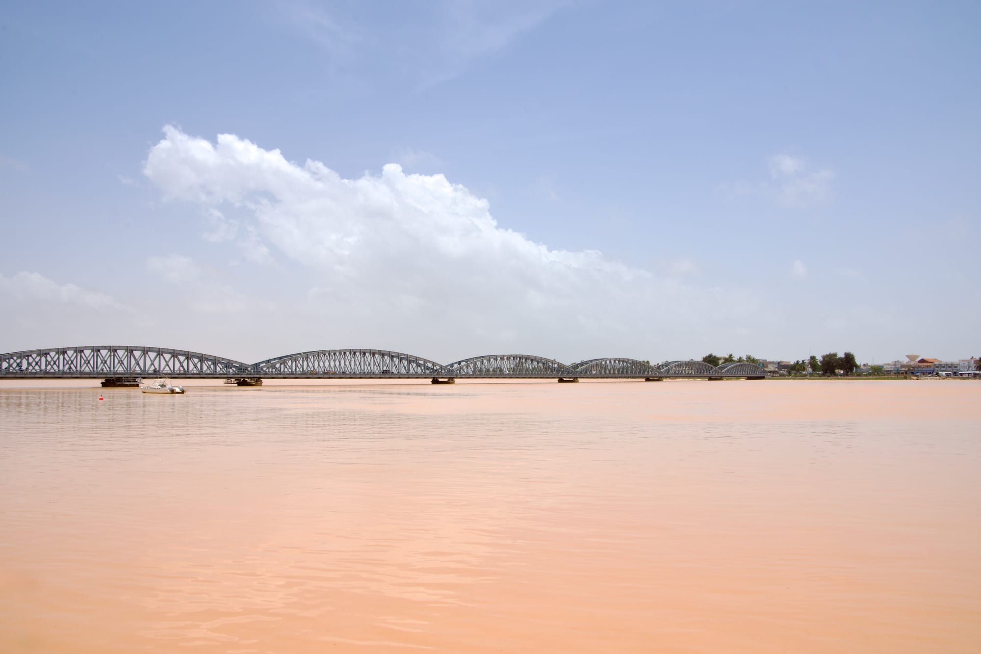 A long grey steel bridge with arched spans crossing a wide, calm river under a pale blue sky