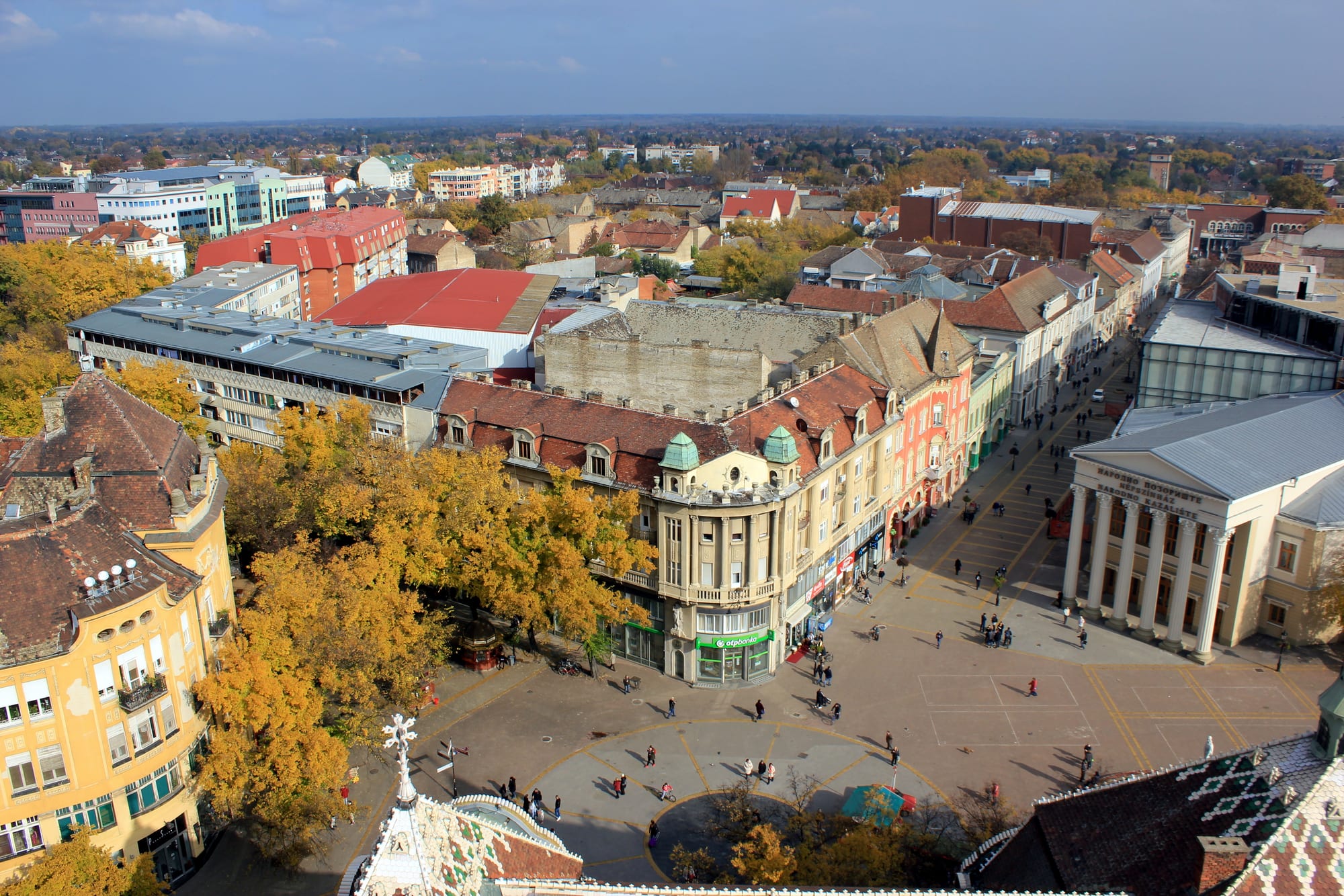 A bird's-eye view over a pedestrian square with a colonnaded theatre, ornate old buildings and autumn trees, with a flat city stretching to the horizon