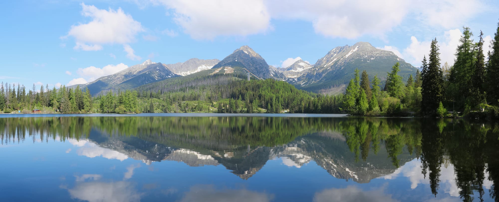 Rocky peaks with patches of snow reflected in a still mountain lake ringed by spruce forest under a blue sky