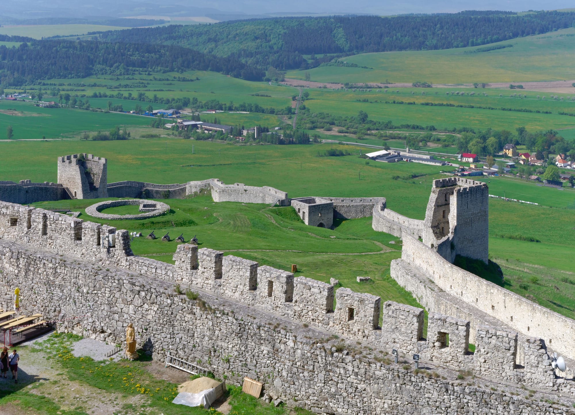 Extensive whitish castle ruins spread along a high ridge above green fields, with hills fading into the distance