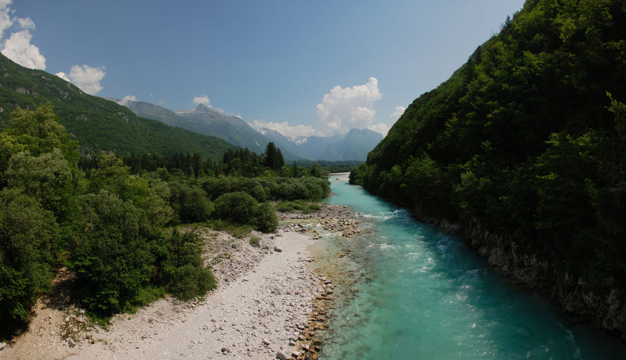 A clear turquoise river running over white gravel between steep wooded slopes, with grey mountains in the distance