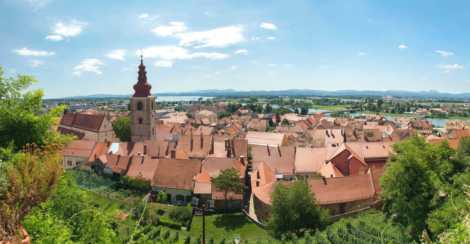 Red-tiled roofs of an old town with a baroque church tower, a river and a lake beyond and hills on the horizon