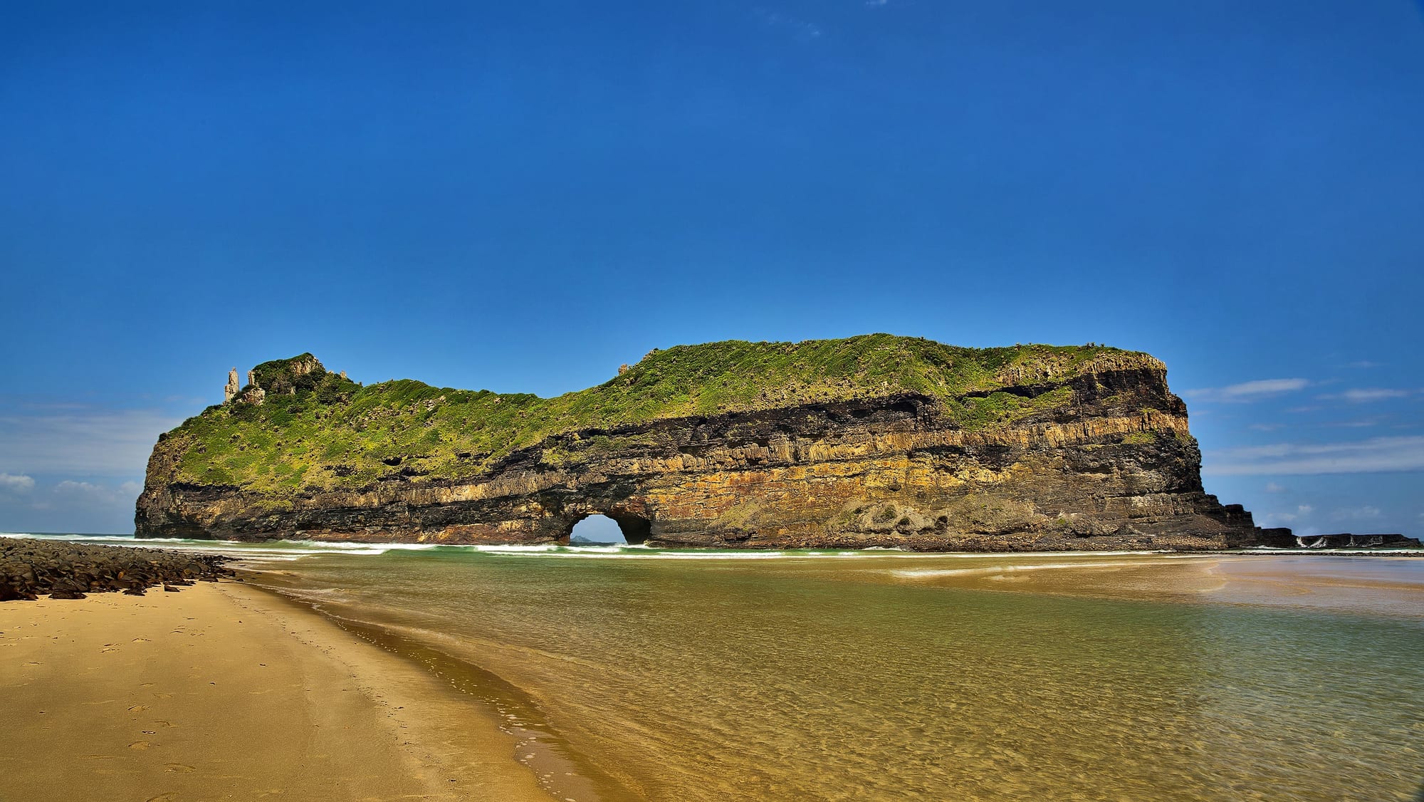 A long green-topped rock island with an arch at sea level, standing off a sandy river mouth under a deep blue sky