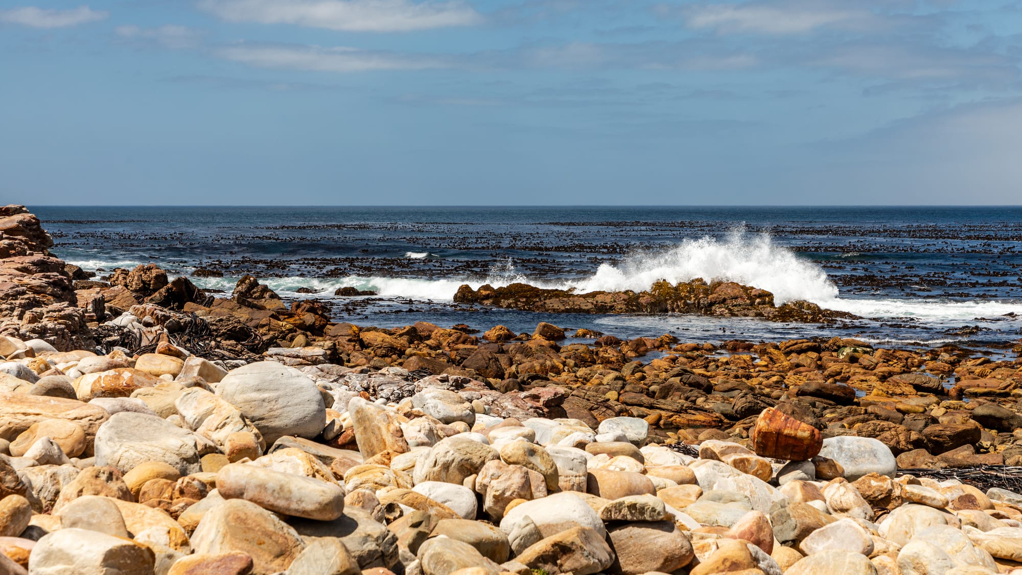 Waves breaking on dark rocks and a shore of rounded pale boulders under a blue sky on the open Atlantic coast