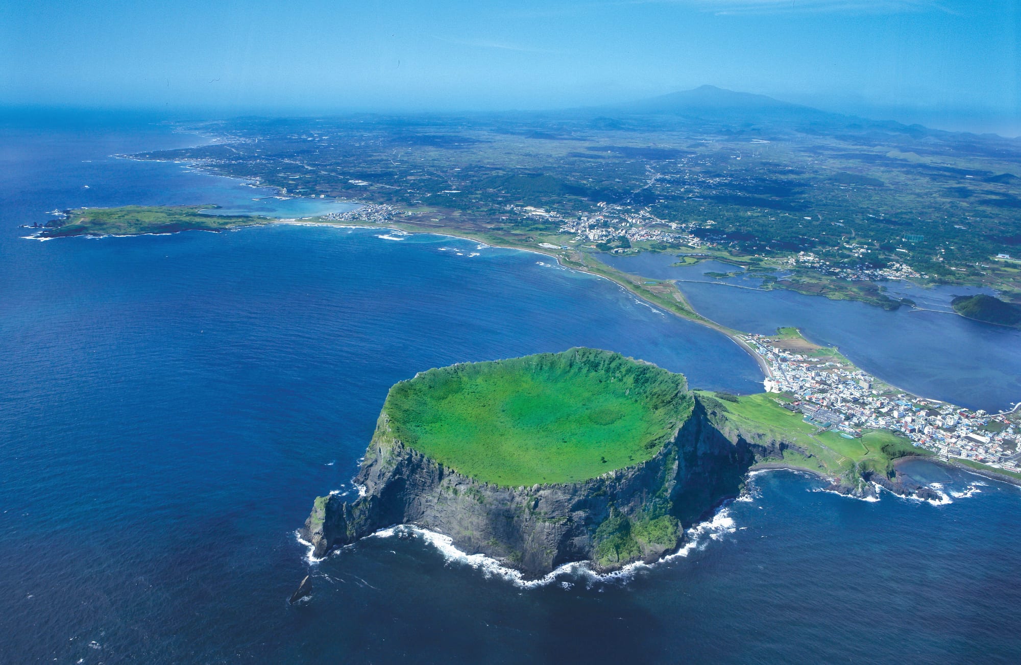 Aerial view of a green, bowl-shaped crater on a sea cliff, joined to a coastal town, with a large mountain in the haze behind