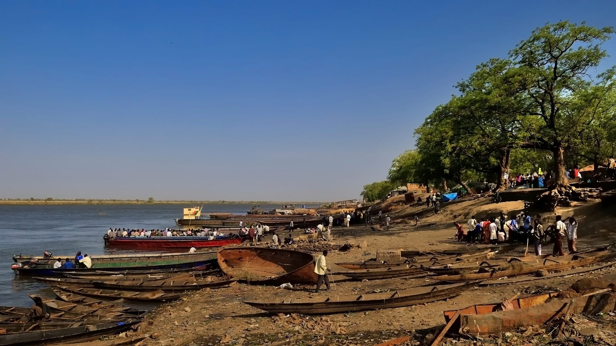 Wooden boats pulled up on a sandy riverbank under shade trees, with passenger boats on the wide Nile beyond