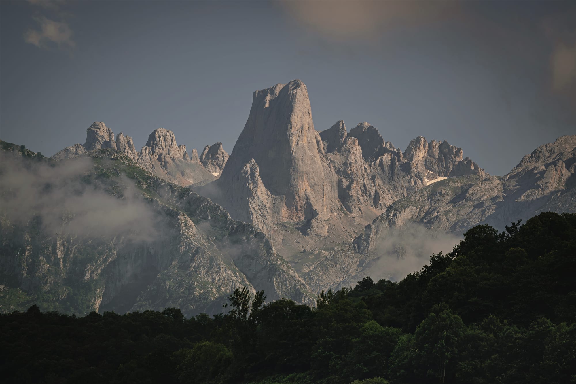 A sheer tower of pale limestone rising above misty ridges and a dark band of forest in the foreground