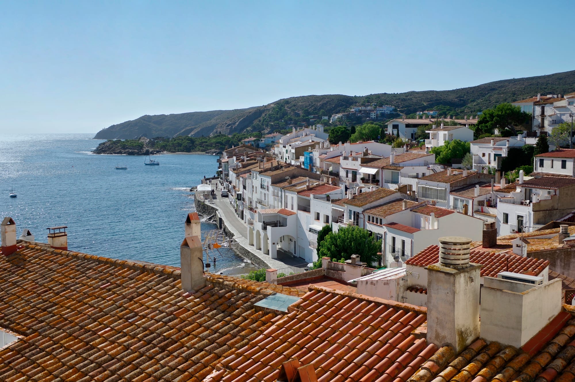 Terracotta rooftops and white houses along a seafront promenade, with a calm blue bay and green headlands beyond