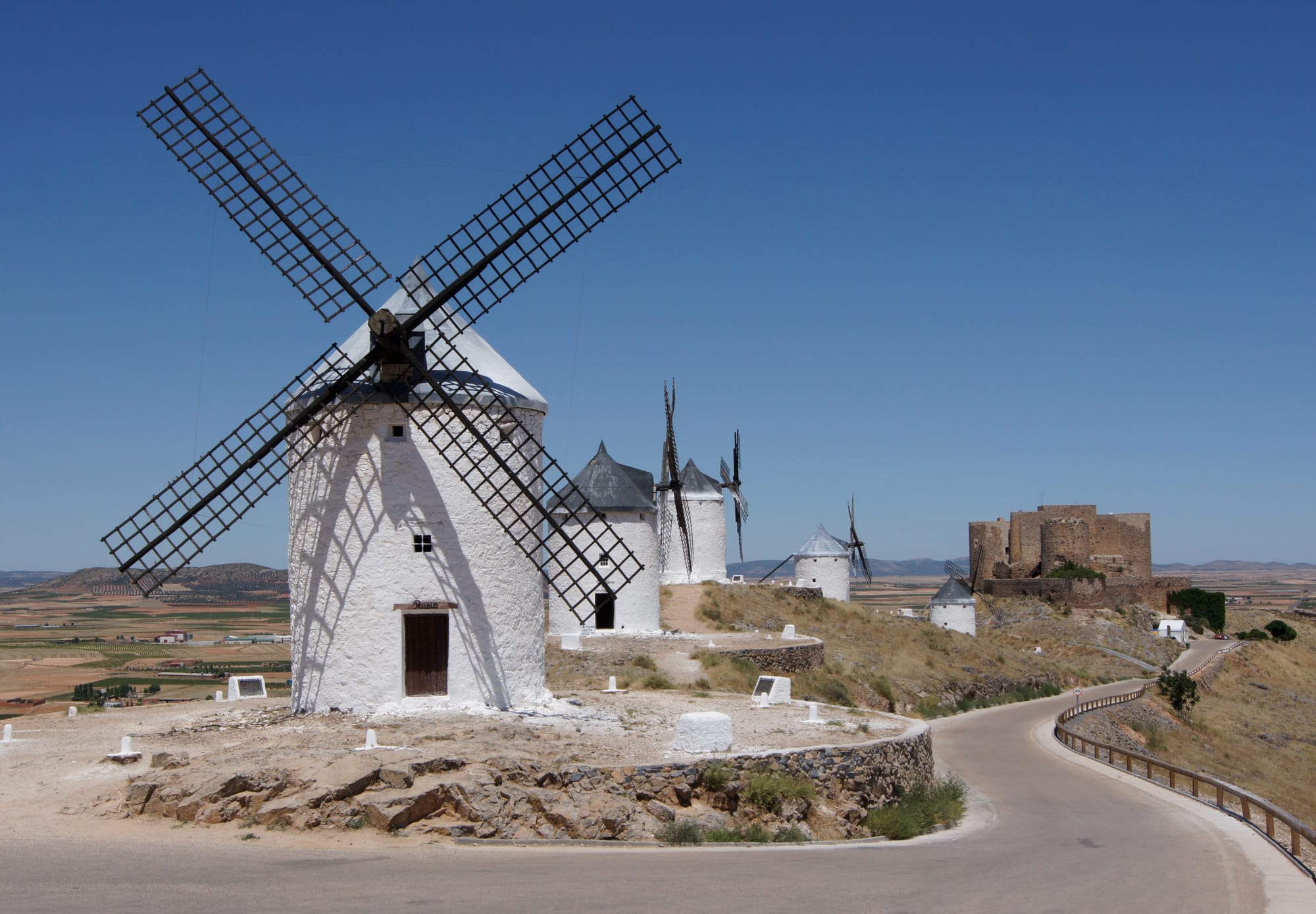 White windmills with black sails on a dry ridge above farmland, with a stone castle at the end of the ridge