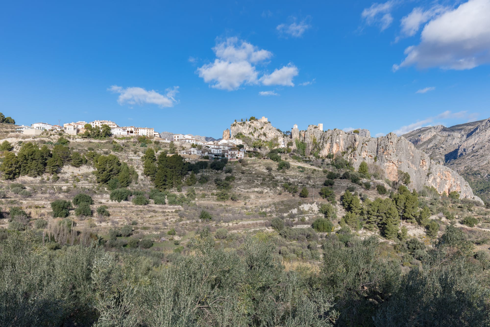 A white hill village and castle ruins on grey limestone crags above terraced slopes of pines and olive trees