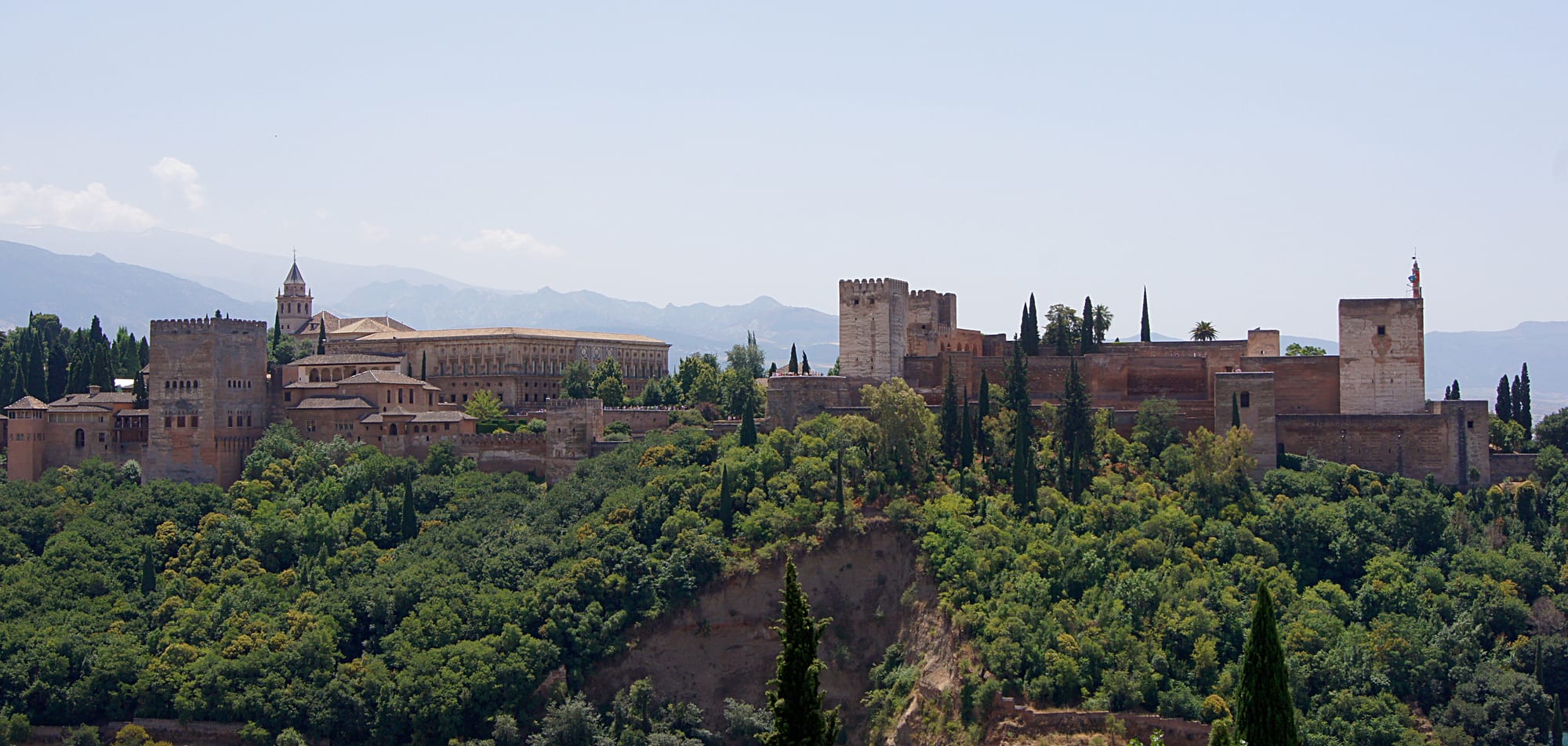 A long hilltop palace-fortress of reddish walls and towers above a city, with snow-streaked mountains rising behind