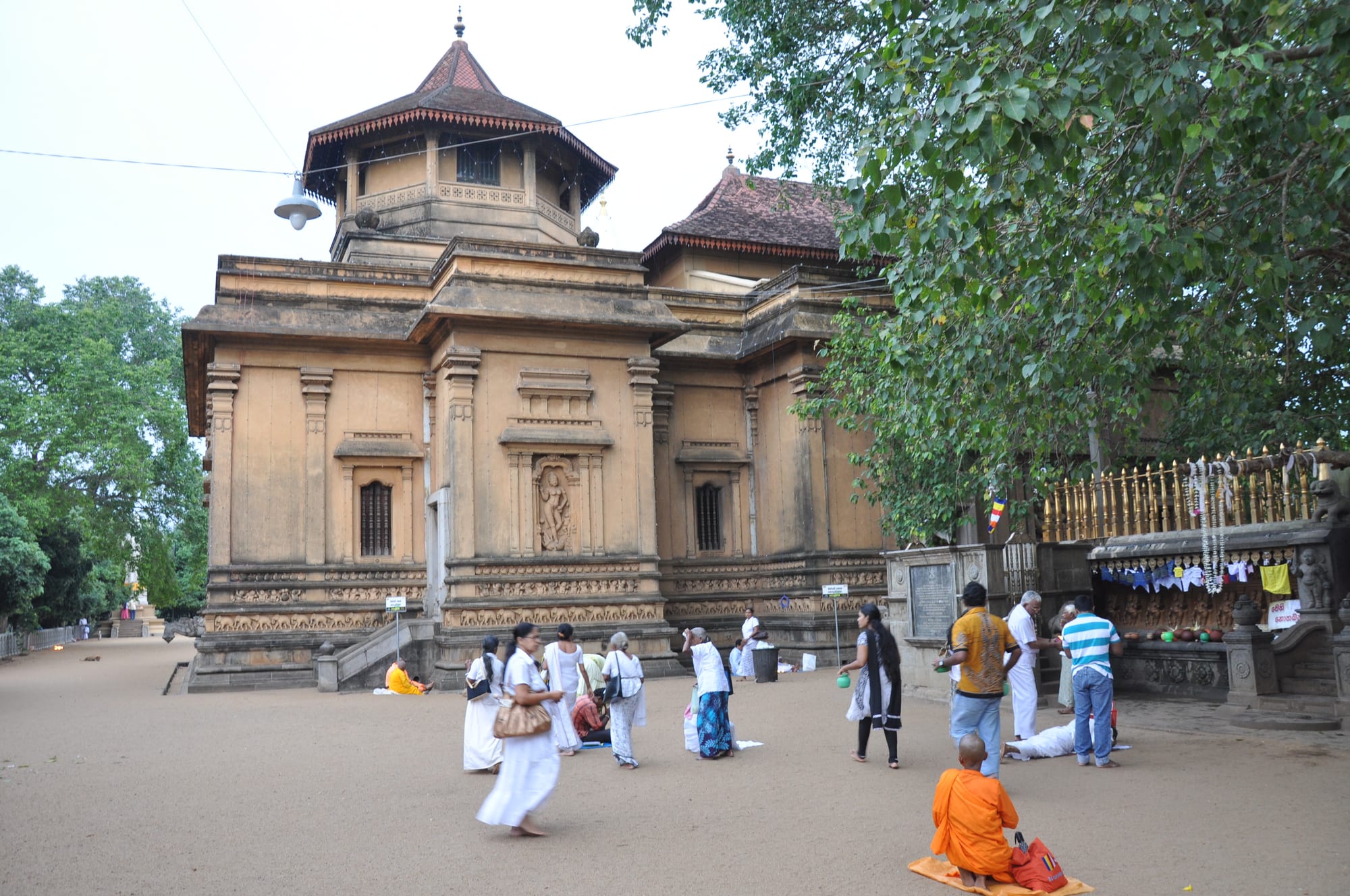 Ochre-coloured shrine with tiled pagoda roofs under a large tree, with white-clad worshippers and a monk in the courtyard