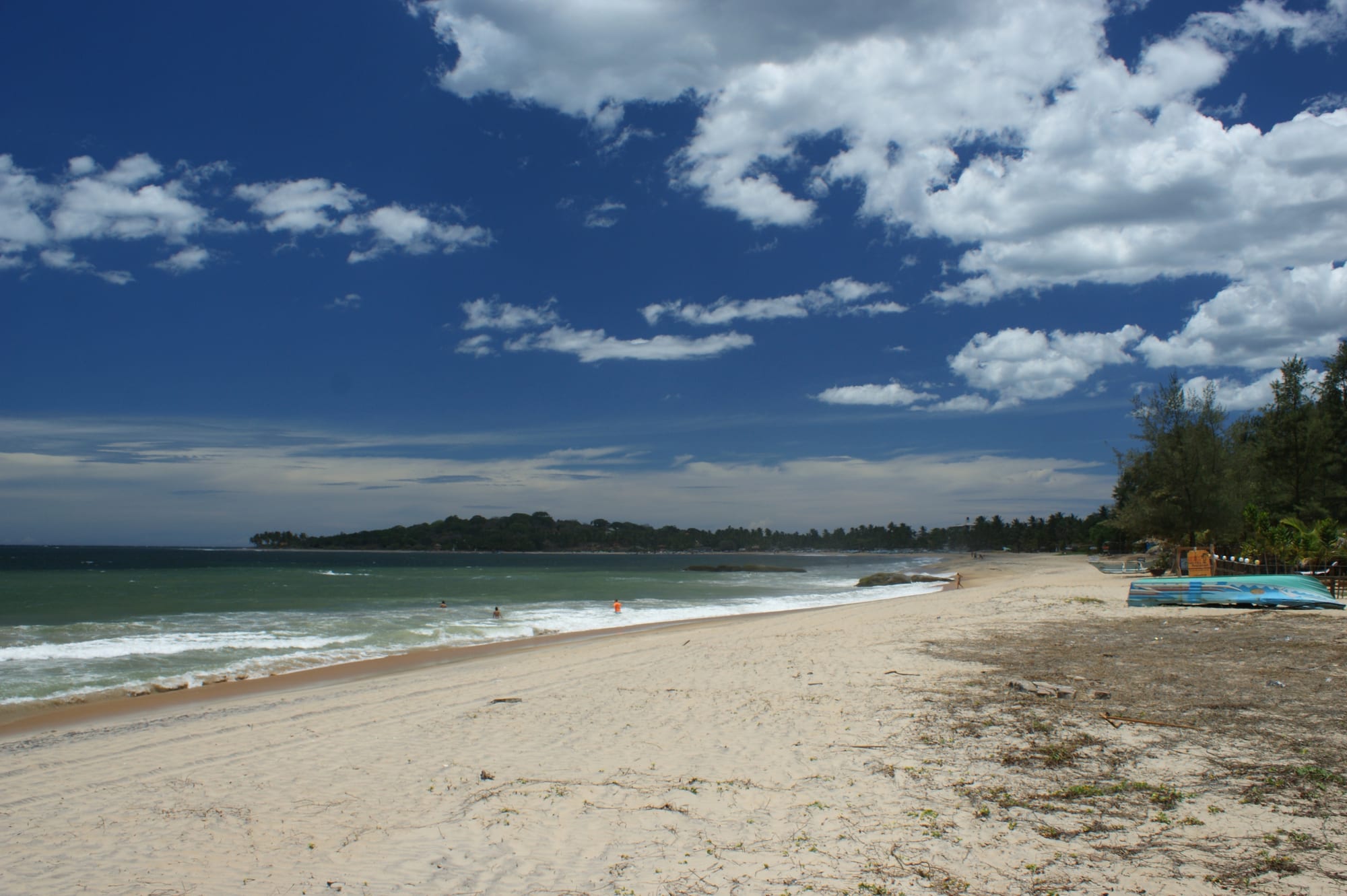 A long sandy beach with small waves under a deep blue sky with white clouds, and a wooded headland in the distance