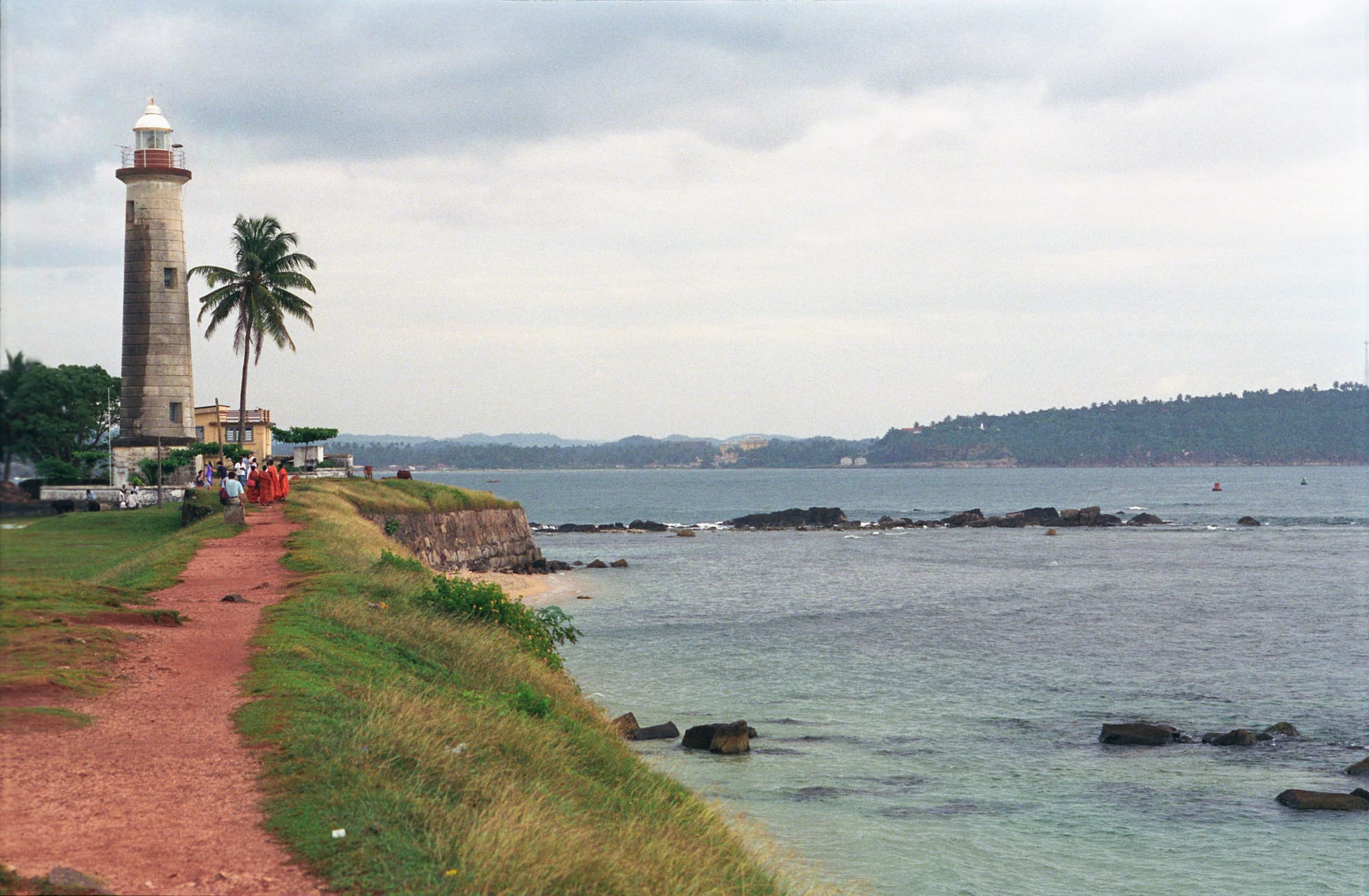 A white lighthouse and palm tree on grassy fort ramparts beside a calm bay, with a green headland across the water