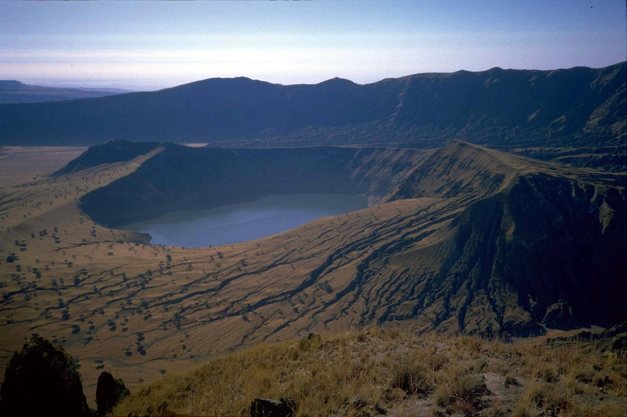 A deep volcanic crater holding a blue lake, ringed by steep dry slopes and ridges under a pale sky