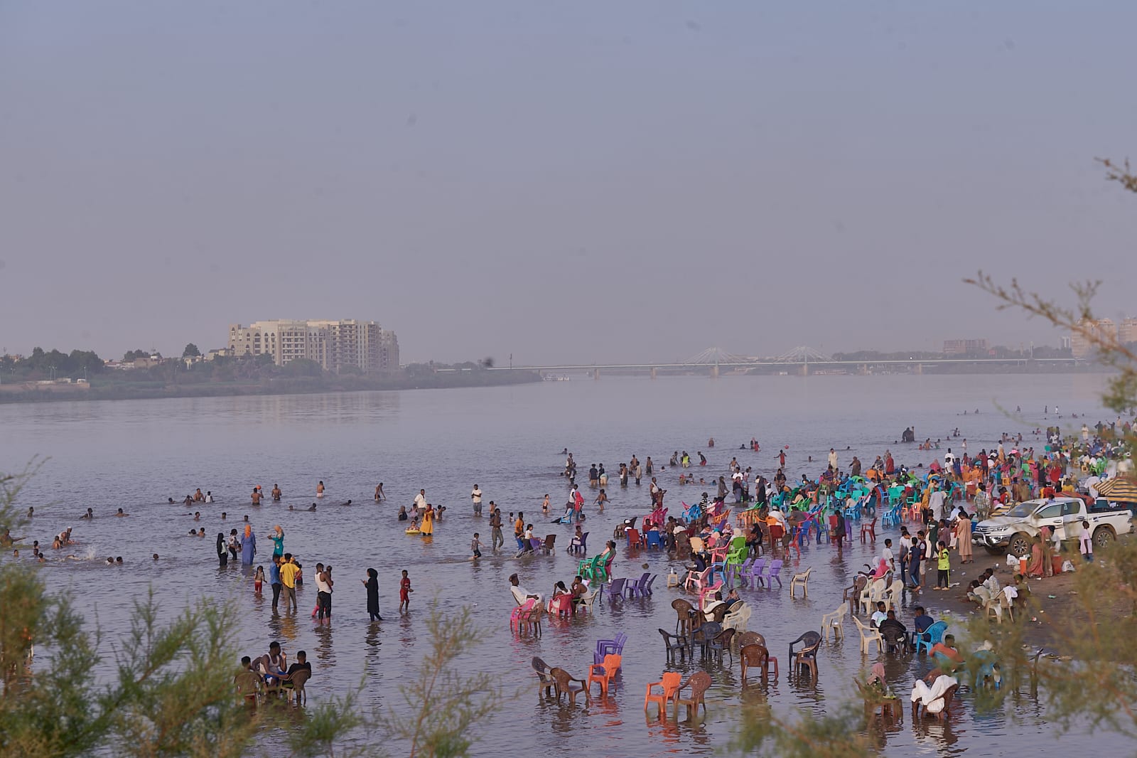 Crowds wading and sitting on plastic chairs in a wide, shallow river, with a long bridge and buildings behind