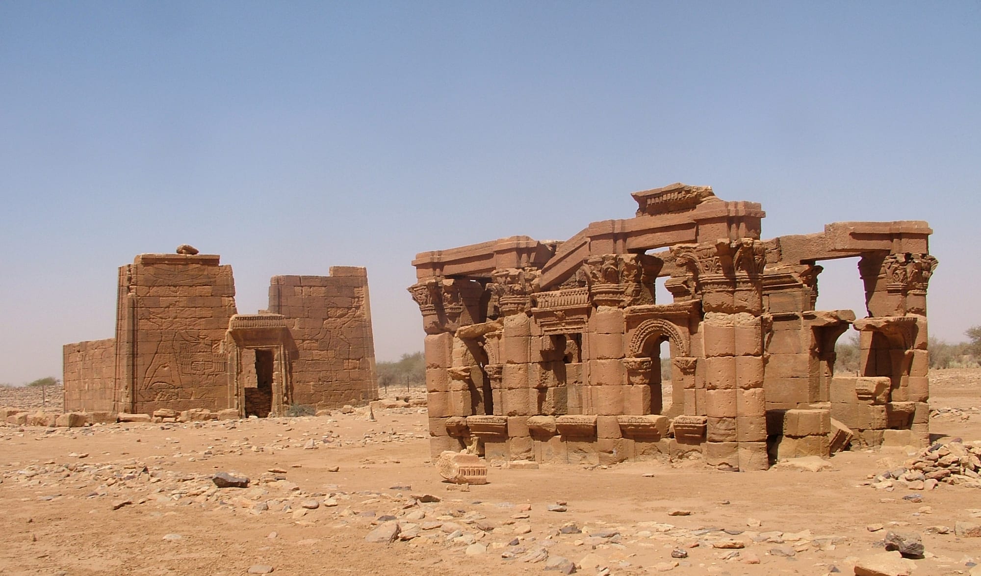 Two sandstone ruins in a stony desert under a clear sky, a columned kiosk with arches on the right and a temple gateway carved with reliefs on the left