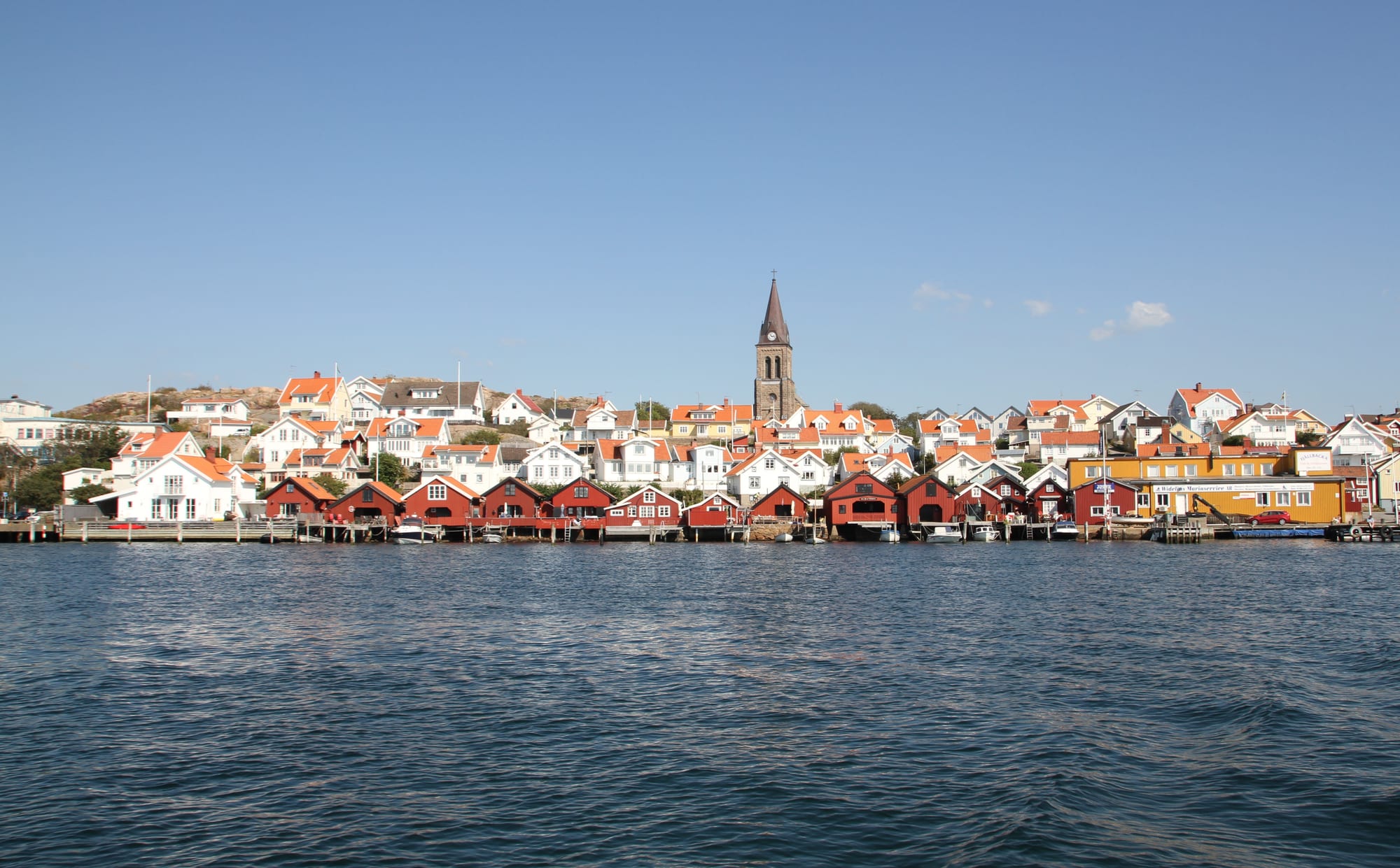 Row of red wooden boathouses along the water in front of white houses and a church spire under a clear blue sky
