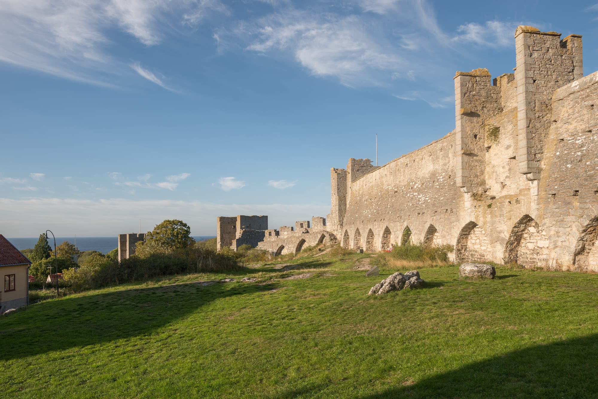 Long medieval stone wall with square towers and arches beside a green lawn, the sea visible in the distance