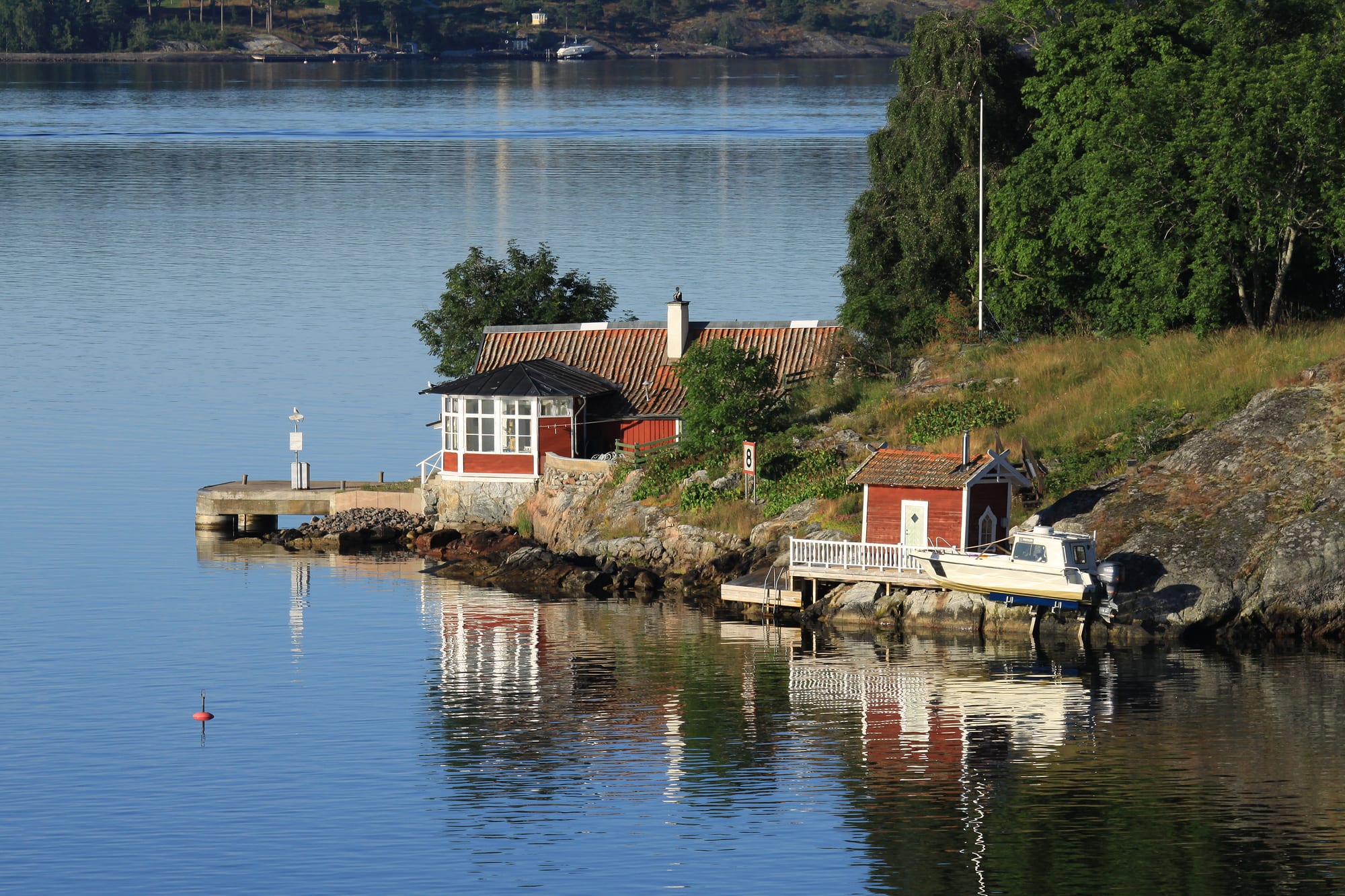 Red wooden cottage and boathouse on a rocky wooded island shore, reflected in calm blue water