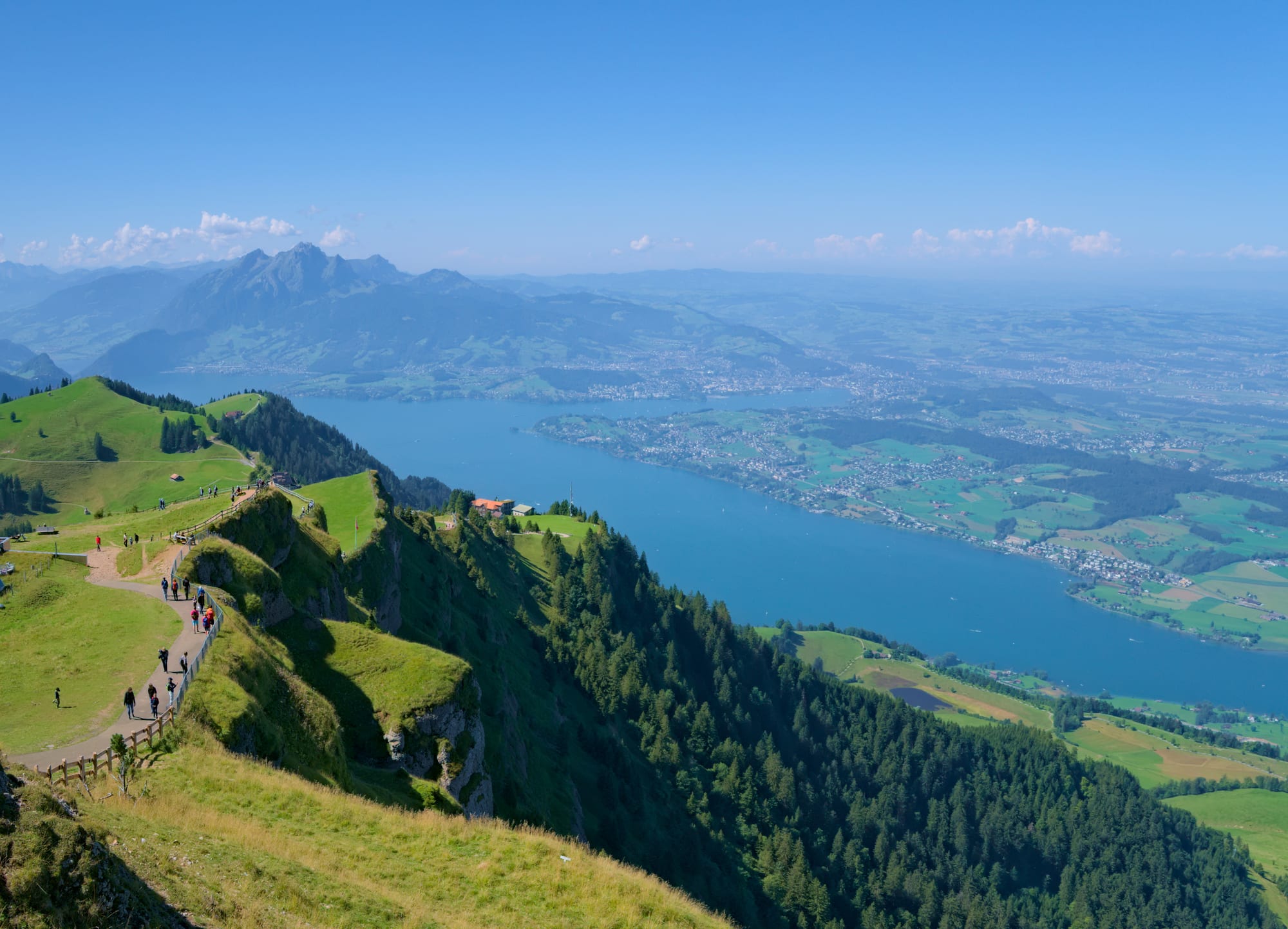 View from a grassy ridge with a walking path down to a long blue lake, a town on the far shore and a jagged mountain behind